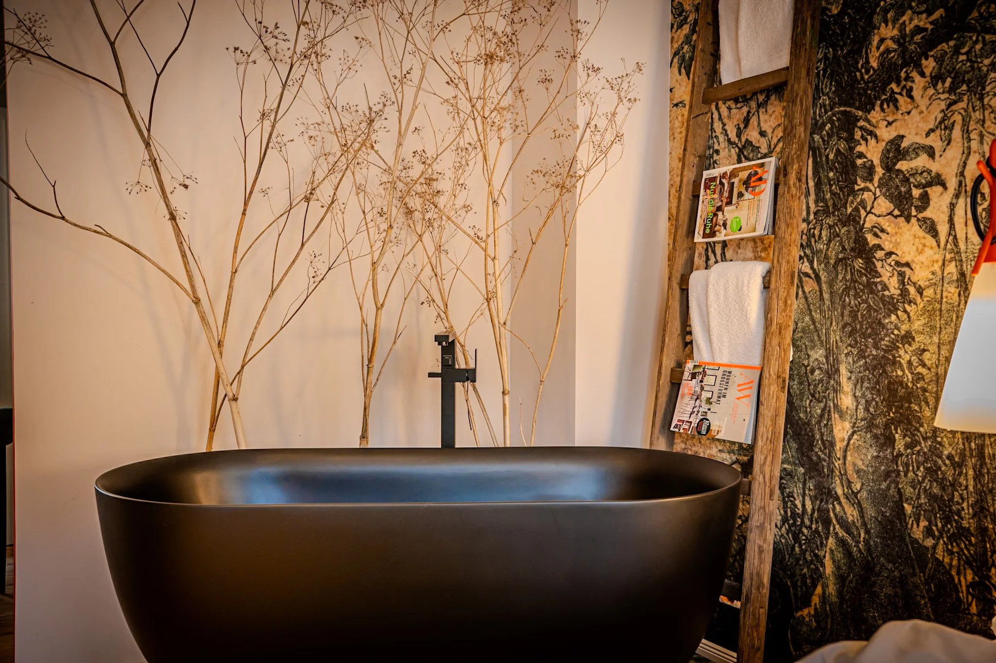 Ein modernes Badezimmer mit schwarfer Badewanne, einer Wand mit aufgehängten Zeitschriften, einem Regal mit weißen Handtüchern und einer Wanddekoration mit einer schwarzen und goldenen Naturmuster