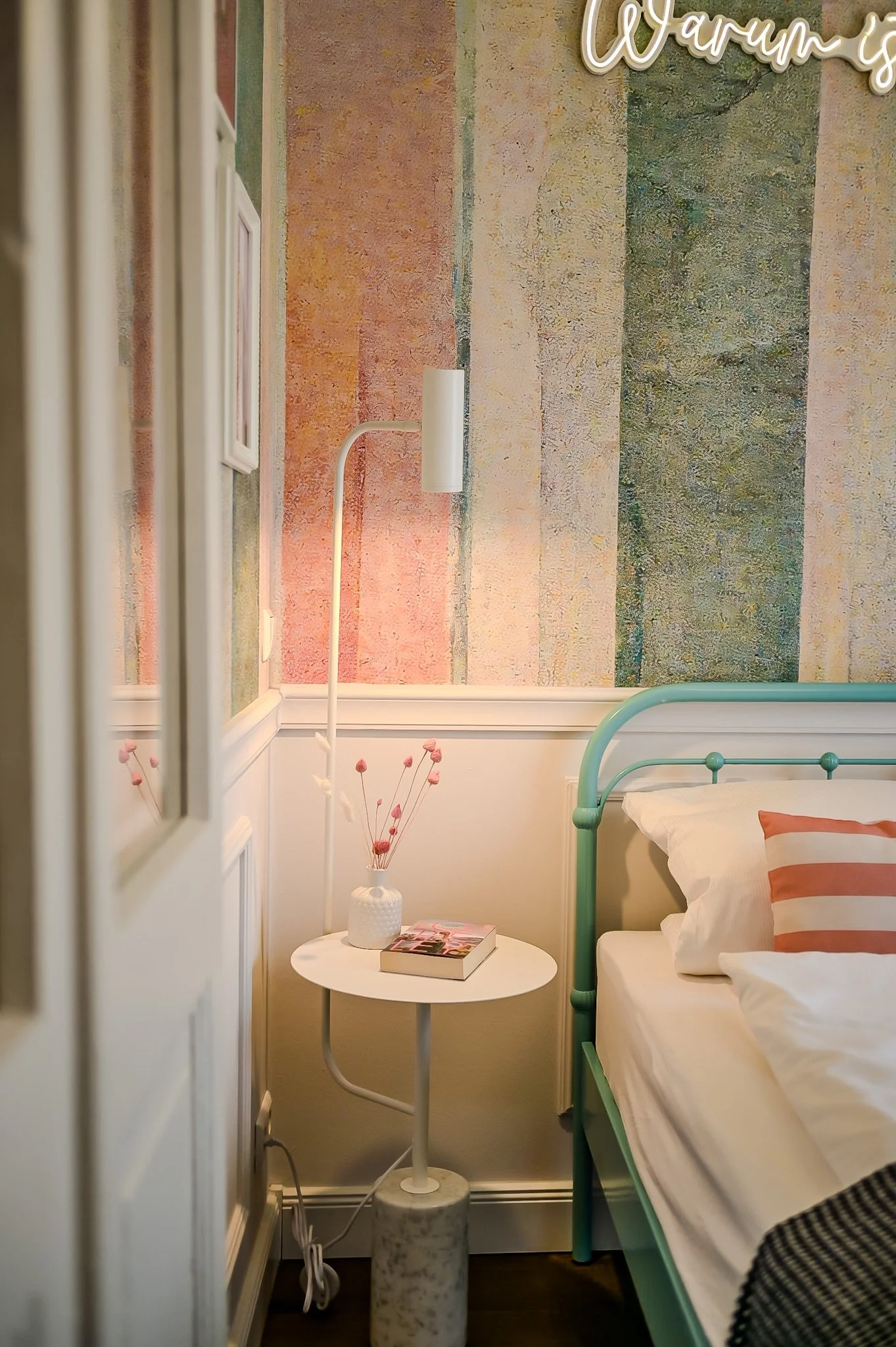 Ein Schlafzimmer mit einem grünen Metallbett, einer weißen Nachttischlampe, einem kleinen runden Beistelltisch mit einer Vase und einem Buch, und einer bunten Wandtapete mit vertikalen Streifen in Pastellfarben, darüber eine leuchtende Wanddekoration
