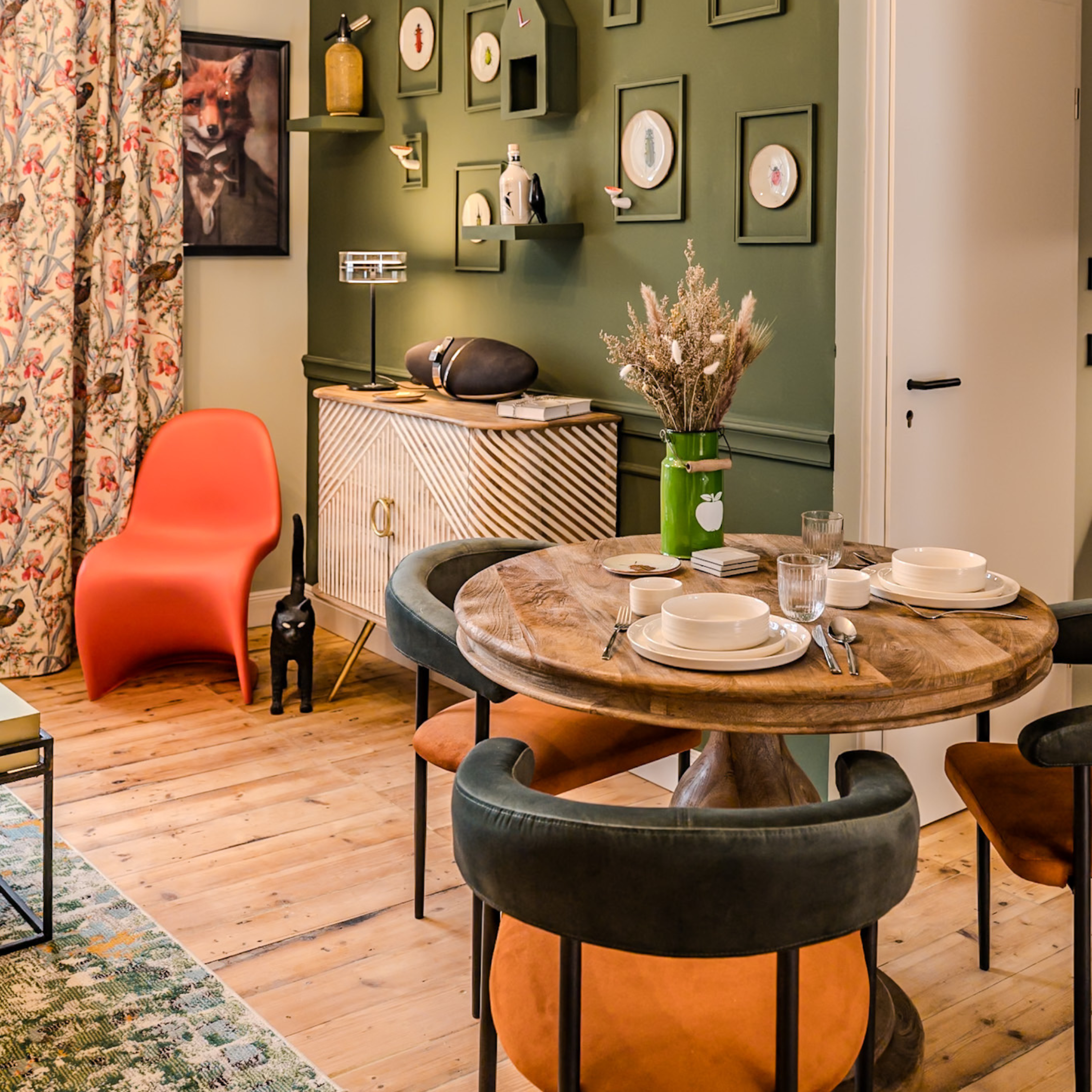 Ein Wohnzimmer mit einem runden Holztisch, gedeckt mit Geschirr und Besteck, umgeben von verschiedenen Stühlen, einer grünen Wand mit Bildern und Regalen, einem Sideboard, einem Korb mit Trockenblumen und einem schwarzen Katzenskulptur