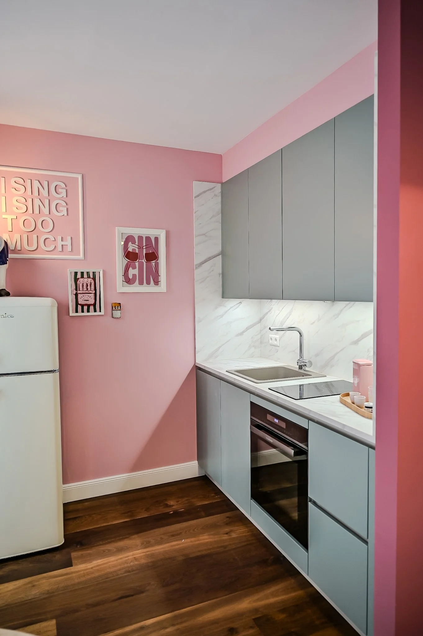 Kleine moderne Küche mit pinken und grauen Schrankfronten, Marmor-Arbeitsplatte, einem elektrischen Herd, einem Spülbecken, dekorativen Wandbildern, Holzfußboden und einer weißen Retro-Kühlschrank.
