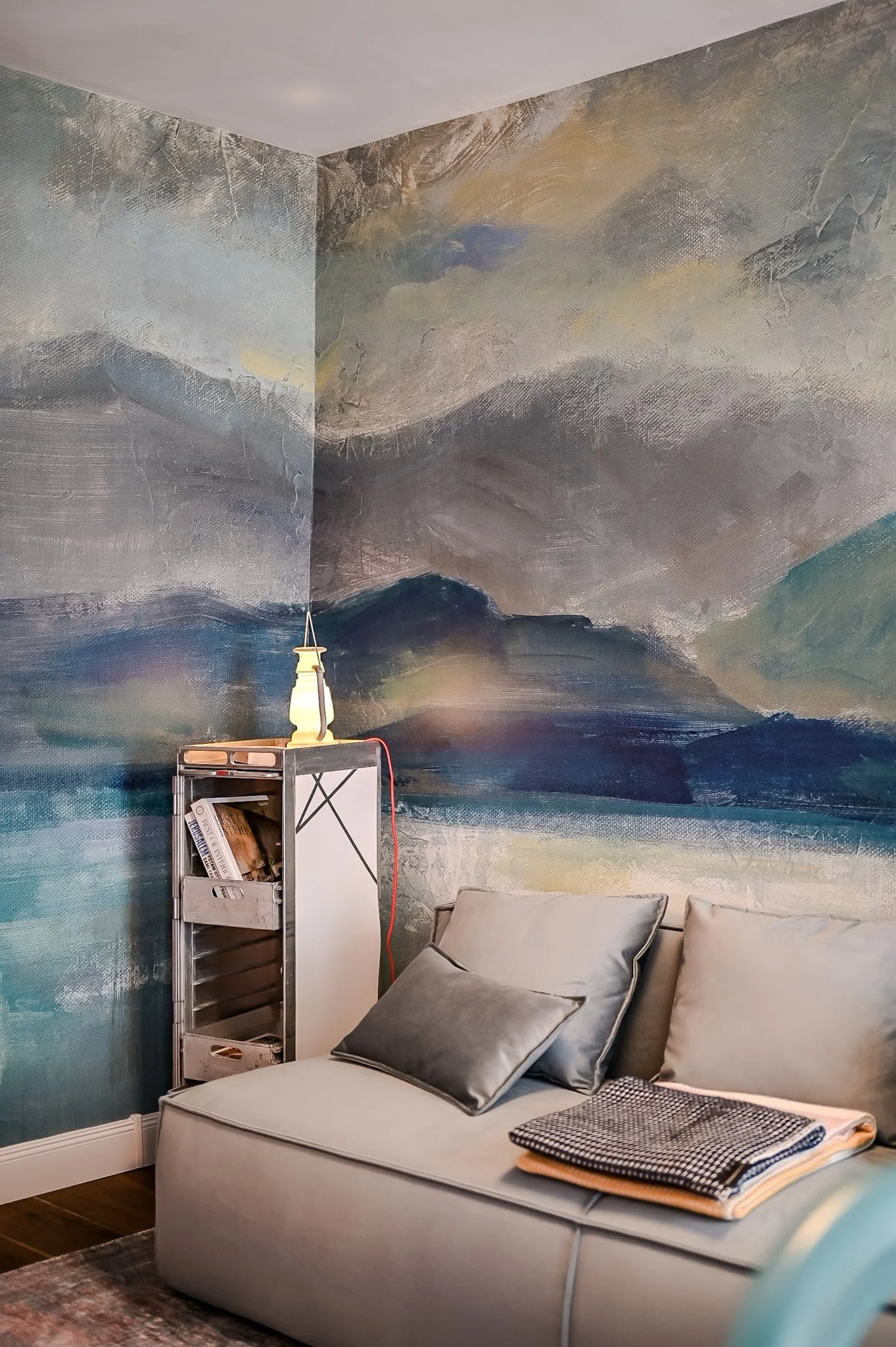 Ein Wohnzimmer mit einer gemalten Wandlandschaft, einem grauen Sofa mit Kissen und einem kleinen Regal, auf dem Bücher liegen, sowie einer Tischlampe auf dem Regal.