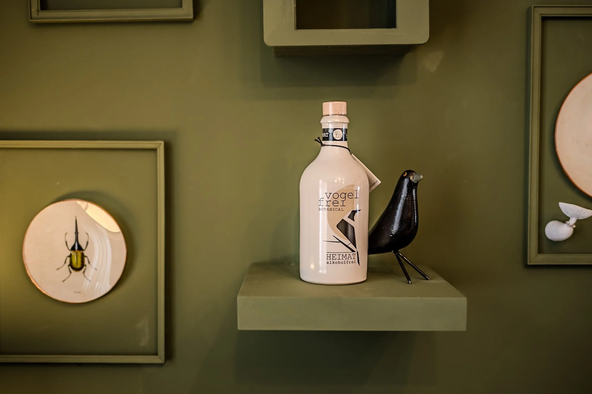 Deko-Objekte auf einer grünen Wand, darunter eine weiße Flasche mit der Aufschrift 'Vogel Frei Botanical Heimat' und ein schwarzer Vogel aus Metall auf einer kleinen grünfarbenen Ablage.