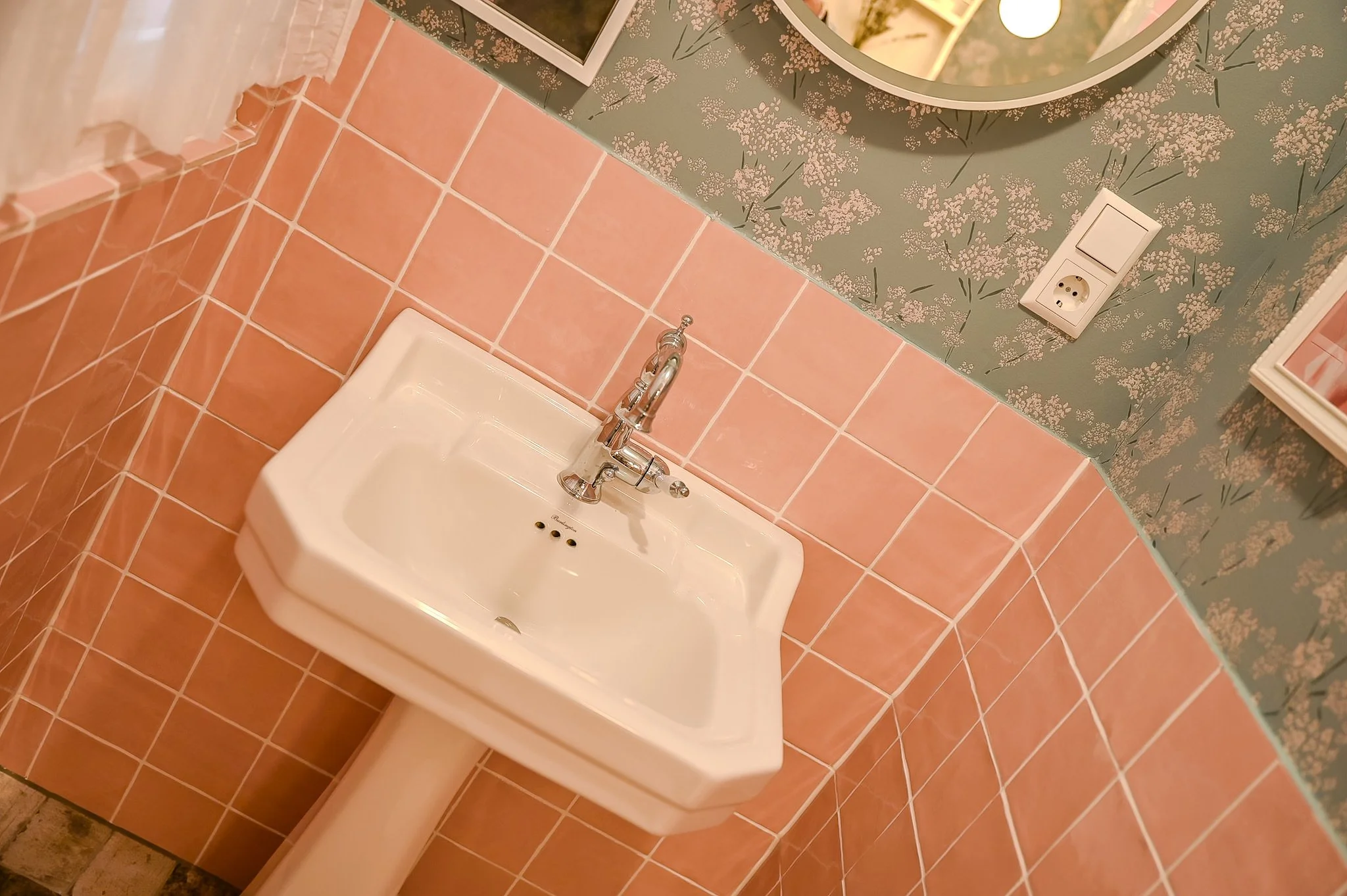 Wandwaschbecken in einem Badezimmer mit rosa Fliesen und floral gemusterter Tapete, eine Spiegel und eine Stromquelle sind sichtbar.
