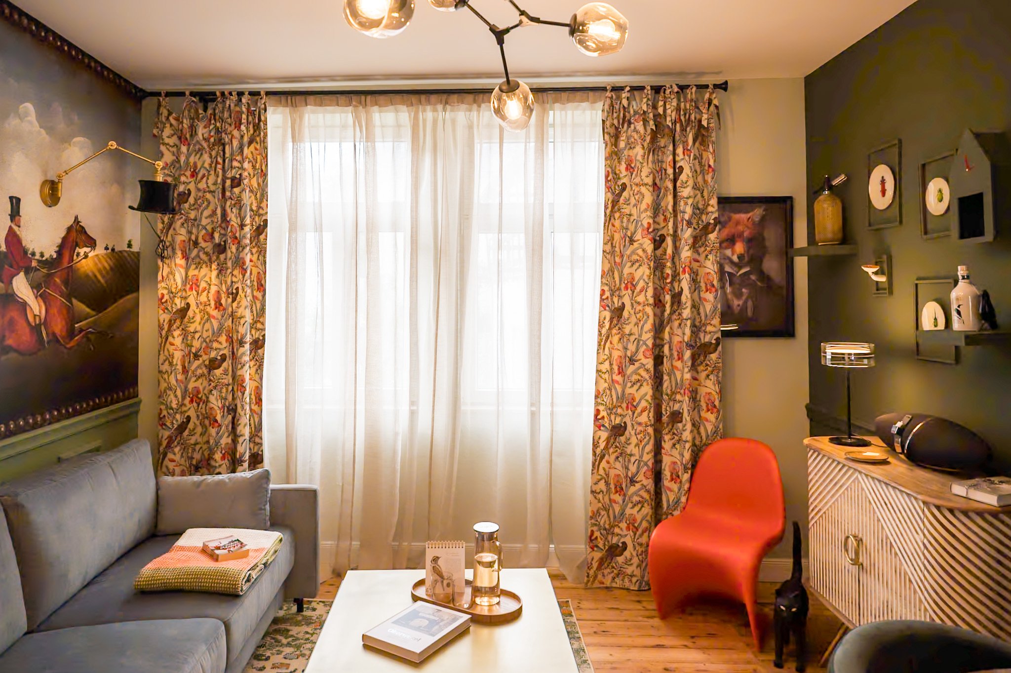 Wohnzimmer mit grauem Sofa, roten Stuhl, Couchtisch mit Büchern und Dekorationen, Wandbildern und Regalen, dicken Vorhängen vor einem großen Fenster, schwarzer Katze.