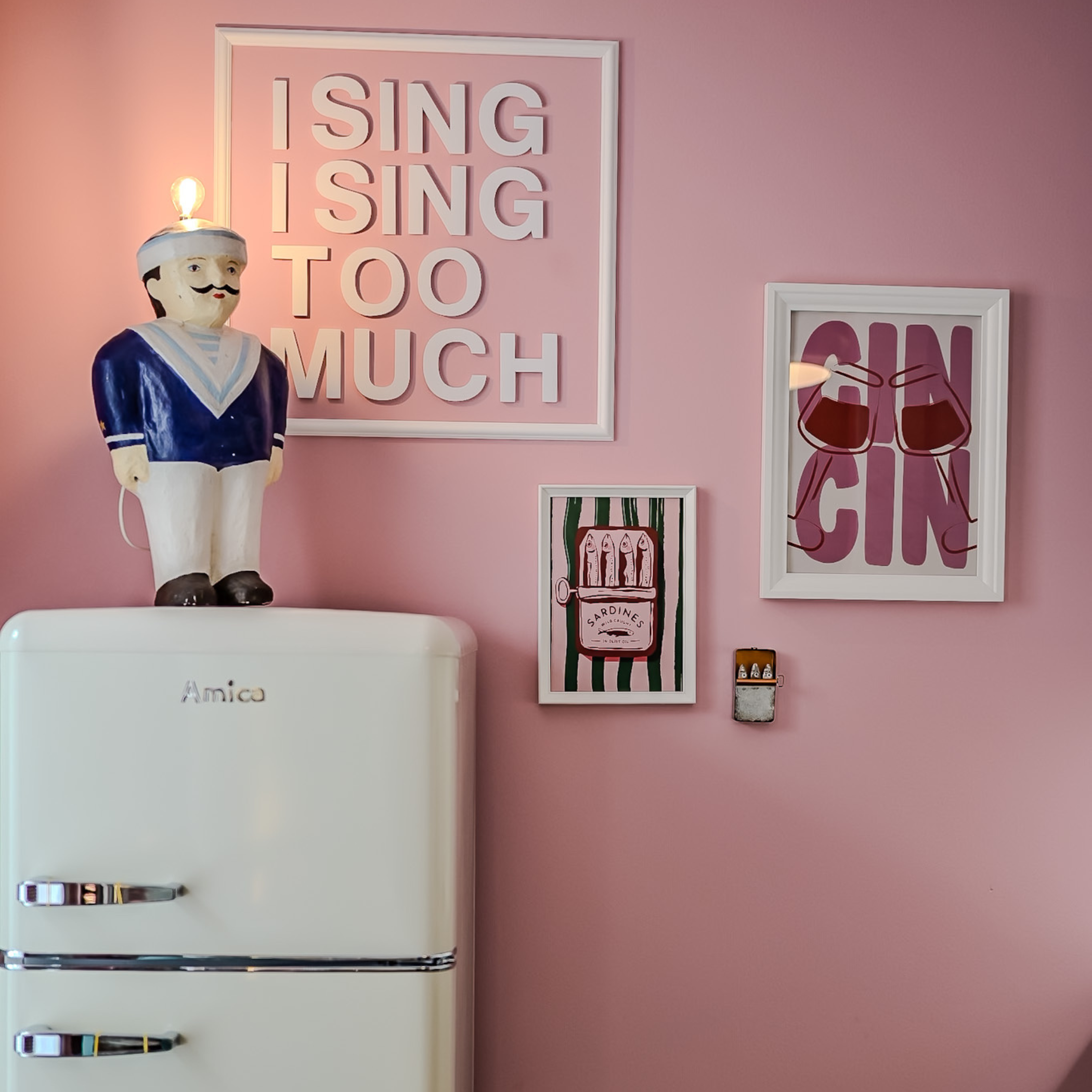 Eine rosa Wand mit vier Bildern und einer Figur an einem alten Kühlschrank. Das größte Bild sagt ‚I SING SING TOO MUCH‘. Ein kleineres Bild zeigt Sardinen in einer Dose. Ein weiteres Bild zeigt zwei Weingläser. Die Figur ist ein kleiner Mann in einer
