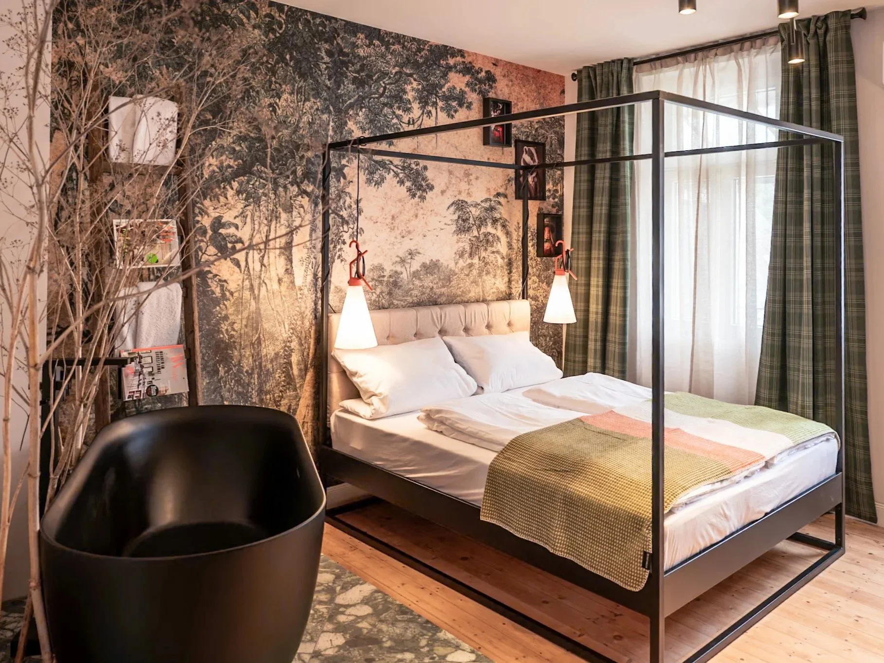 Schlafzimmer mit Himmelbett, Wandmalerei mit Naturmotiv, schwarzer Sessel, große Fenster mit grünen karierten Vorhängen, Holzfußboden, stehlampen über dem Bett, Regale mit Dekorationen an der Wand.