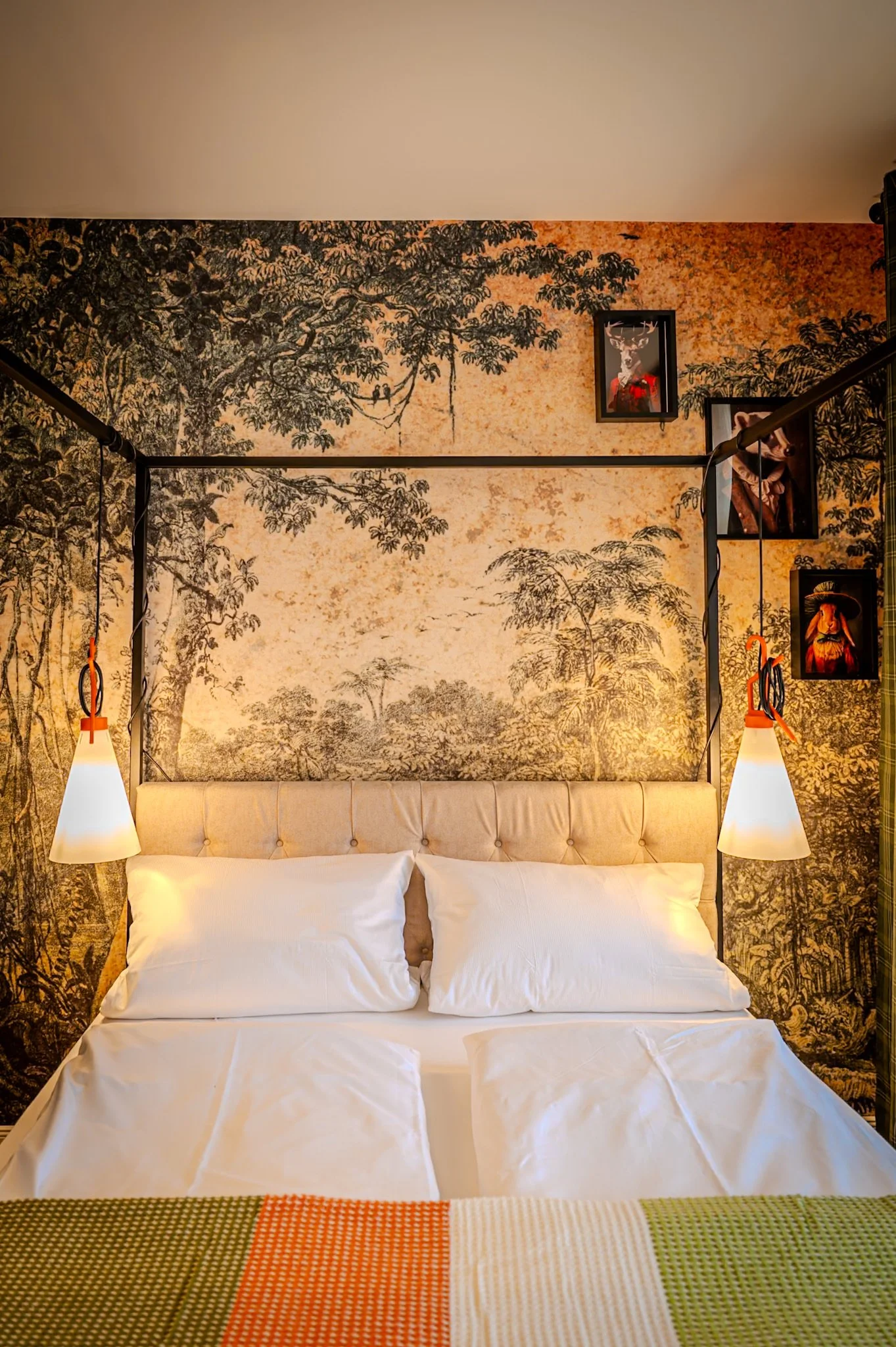 Bett mit weißen Kissen vor einer Wand mit Dschungel-Tapete. Zwei Lampen hängen seitlich am Bett, und es gibt einige Bilder an der Wand.