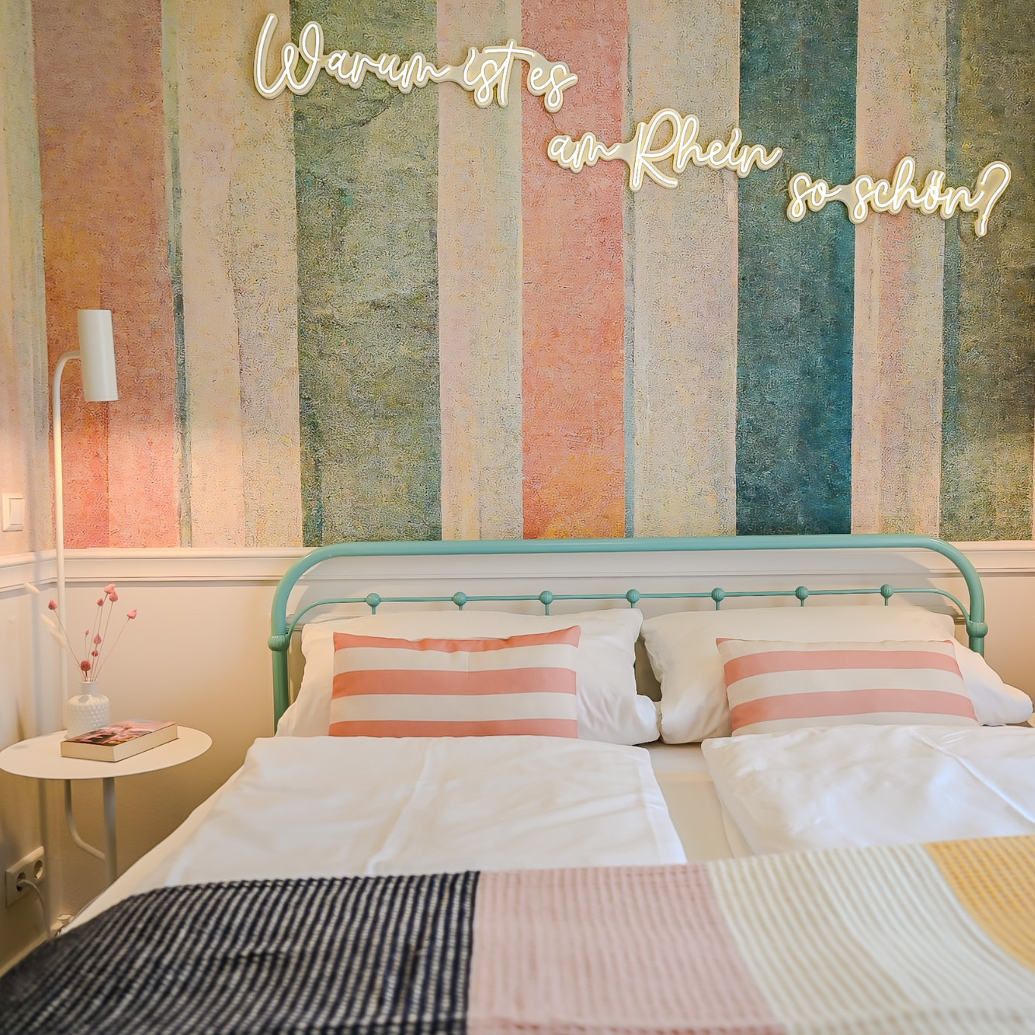 Ein gemütliches Schlafzimmer mit einer blauen Metallkopfleiste, zwei weißen Kissen mit rosa Streifen, einem bunten Patchwork-Bettüberwurf und einem bunten Wandtapete im Retro-Stil. An der Wand hängt eine Lichterkette mit der Aufschrift "Warum ist es 