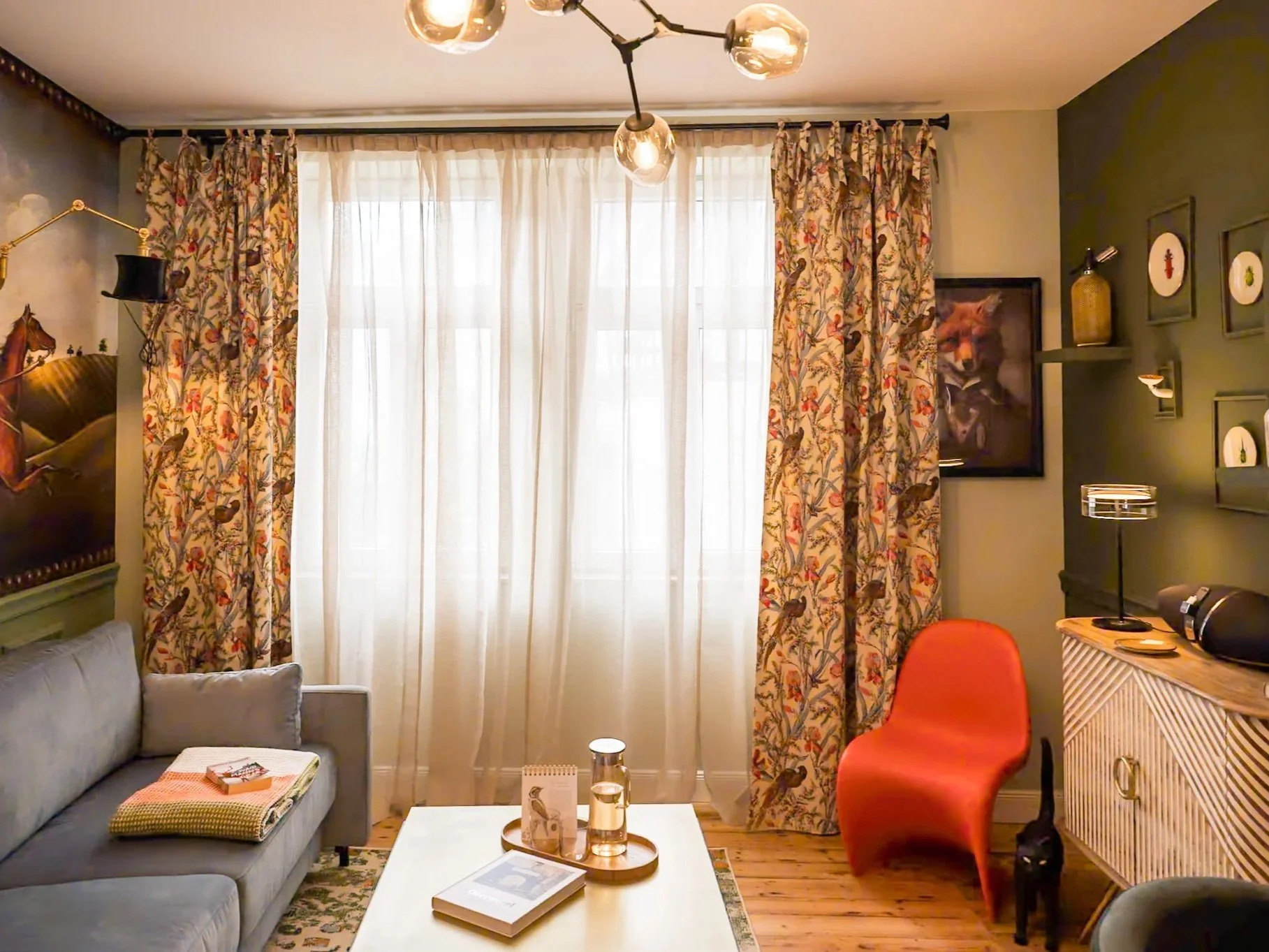 Gemütliches Wohnzimmer mit großen Fenstern, bunten Vorhängen, grauem Sofa, roter Sessel, Couchtisch mit Wasser und Zeitschrift, grüner Wand mit gerahmtem Tierbild, Beleuchtung und Dekorationen.