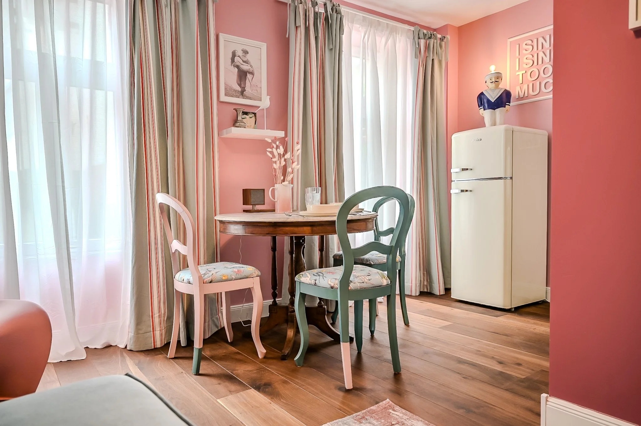 Heller Wohnraum mit pinkfarbenen Wänden, Holzboden, einem runden Esstisch mit vier Stühlen, Vorhängen und einem weißen Kühlschrank im Hintergrund. Es gibt Dekorationen wie Bilder, Skulpturen und einen Stuhl mit gepolstertem Sitz. Auf der Wand liegt e