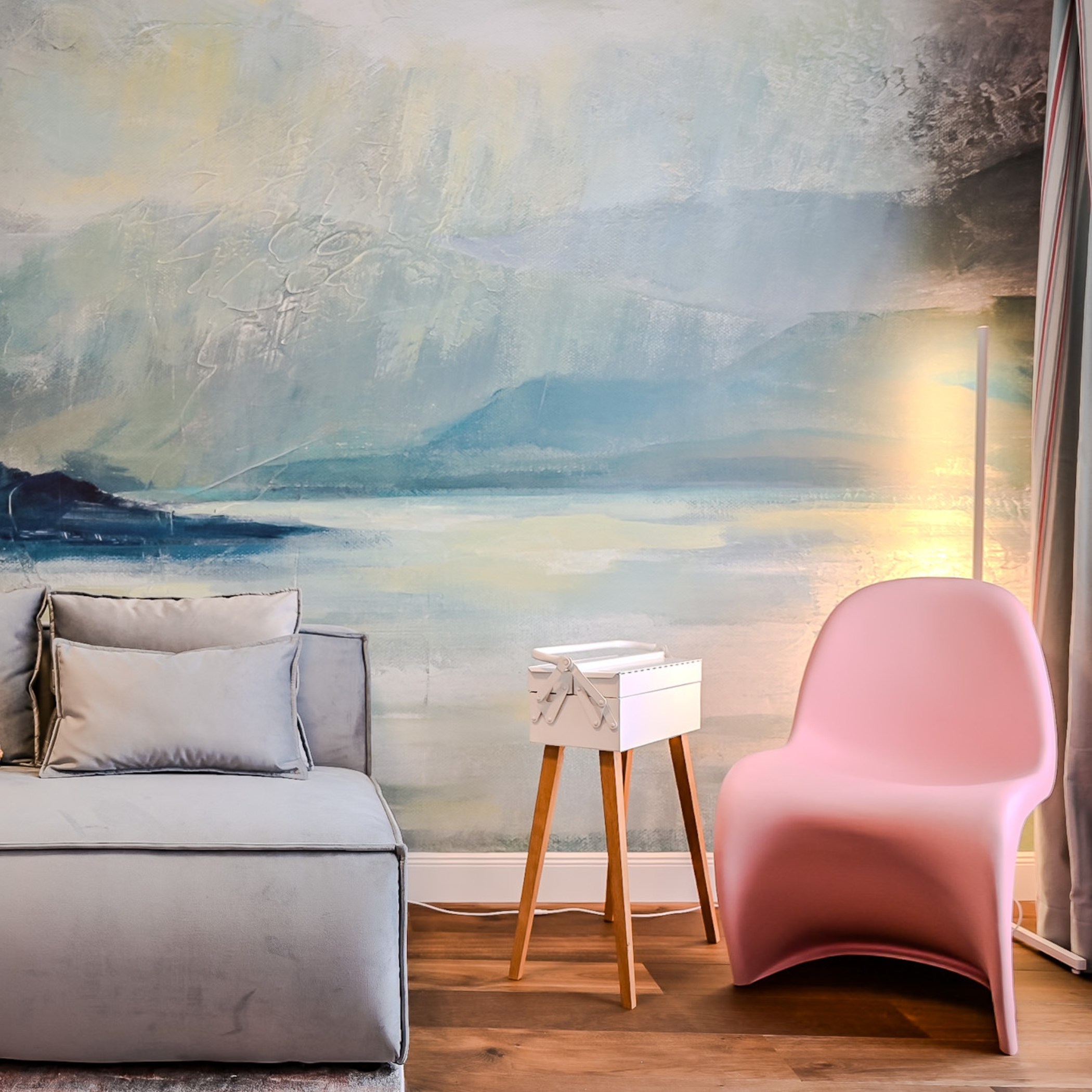 Wohnzimmer mit grau Sofa, pinker Sessel, kleiner weißer Beistelltisch und Wandbild eines Seen- und Bergpanoramas.
