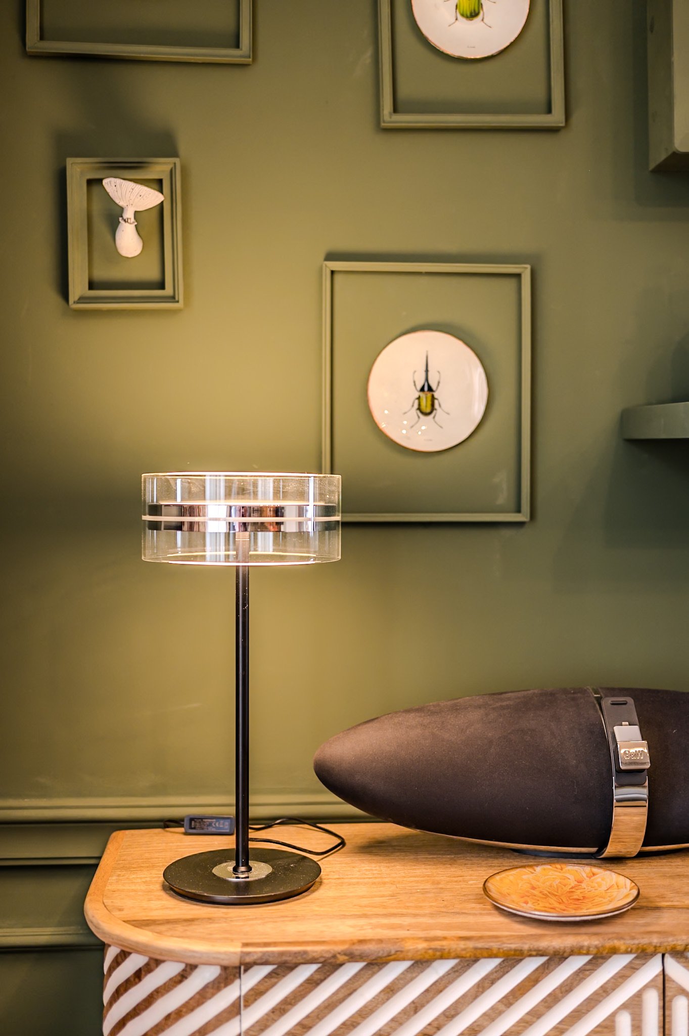 Ein Holztisch mit einem modernen transparenten Tischläufer, einer schwarzen Tischlampe, einem Lautsprecher und einer kleinen Schale. Die Wand im Hintergrund ist grün und hat mehrere aufgekippte Bilder mit Insekten