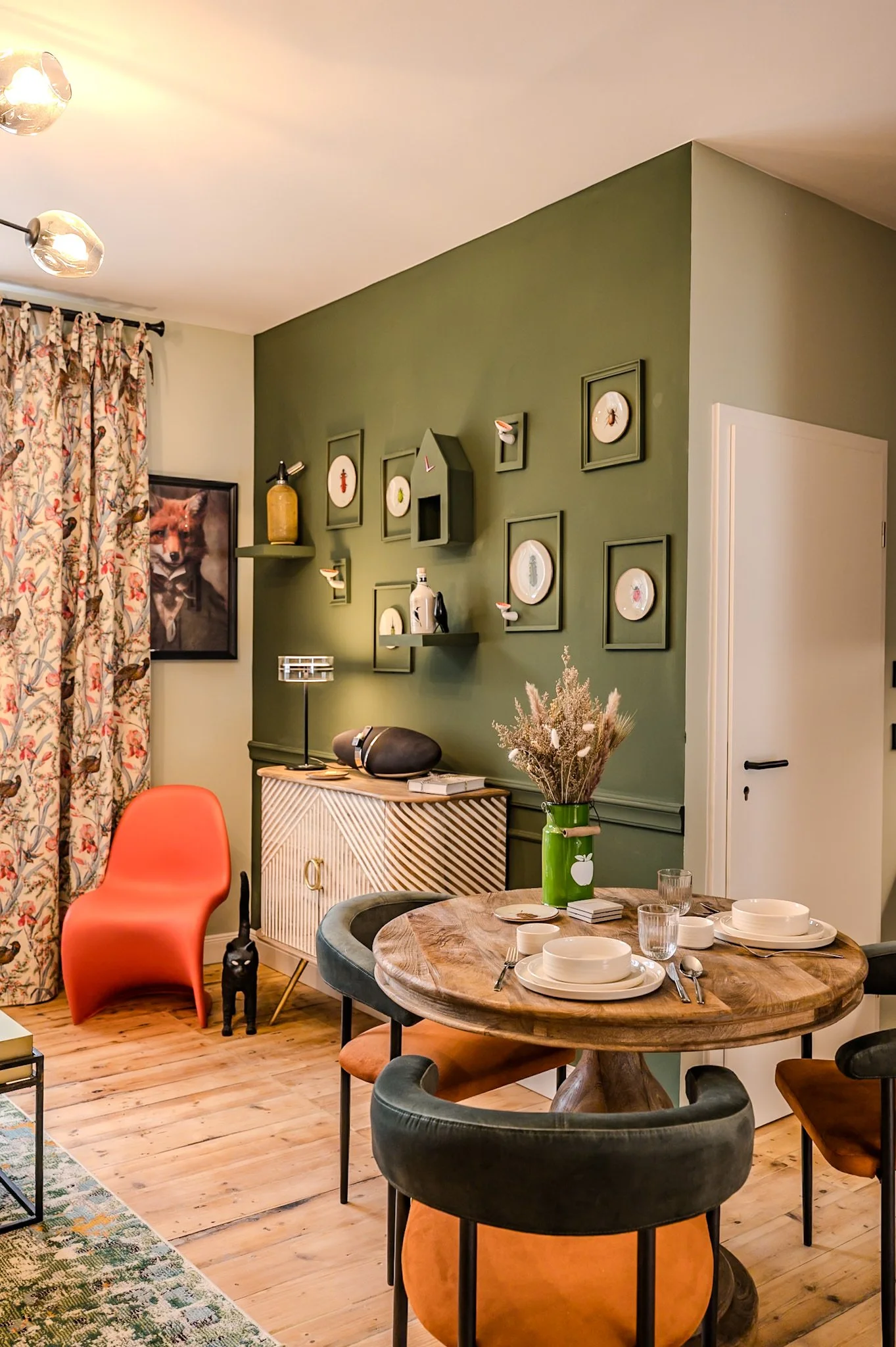 Gemütliches Esszimmer mit grün gestrichener Wand, festlich gedecktem Tisch, blick auf die Wand mit Bilderrahmen und Dekorationen, blumen im grünen Vase, und ein lila Sessel.