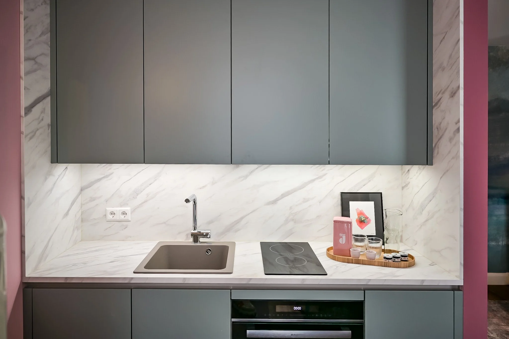 Moderne Küche mit grauen Schränken, weißer Marmor-Arbeitsfläche, einem Einbauspülbecken, einer Herdplatte, einem Wasserglas, einer pinken Thermoskanne, einem Bilderrahmen und einem Holztablett mit Gläsern und kleinen Verpackungen.