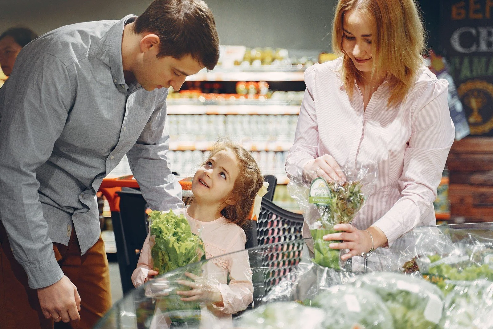 Parents and their young daughter choosing vegetables together at a supermarket, showing how children learn about money by observing everyday choices.