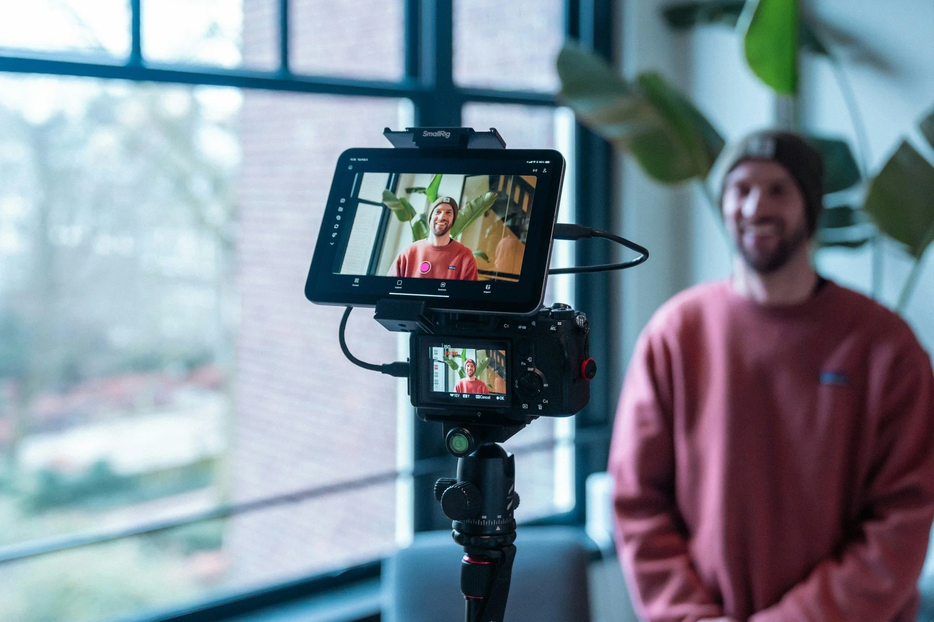A camera on a tripod films a smiling content creator recording an investing video indoors