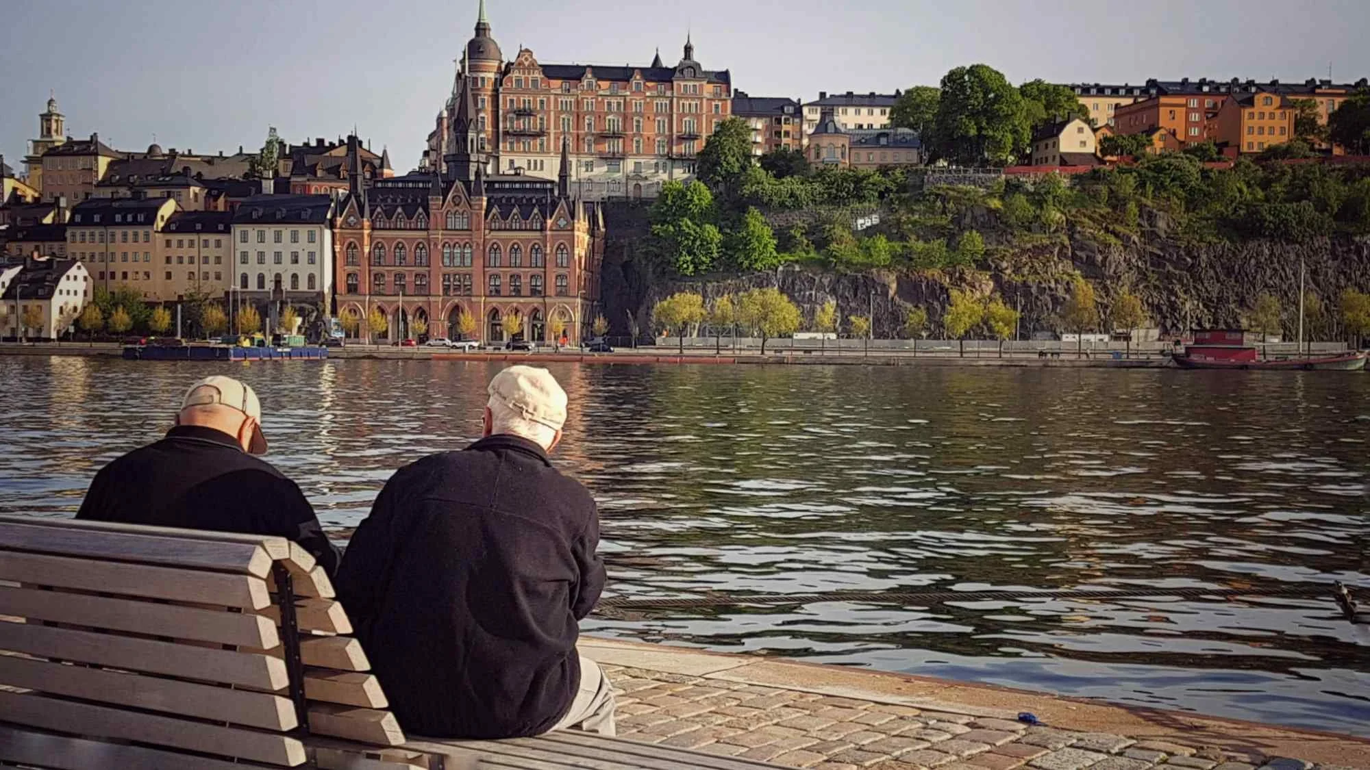Two older friends sitting on a bench by the water in a European city, representing the slow-go years of the retirement spending smile.
