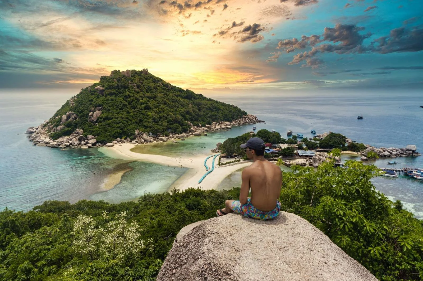 Sunset view over a tropical Thai island peninsula with turquoise water, sandy beach, and a traveler overlooking the bay.