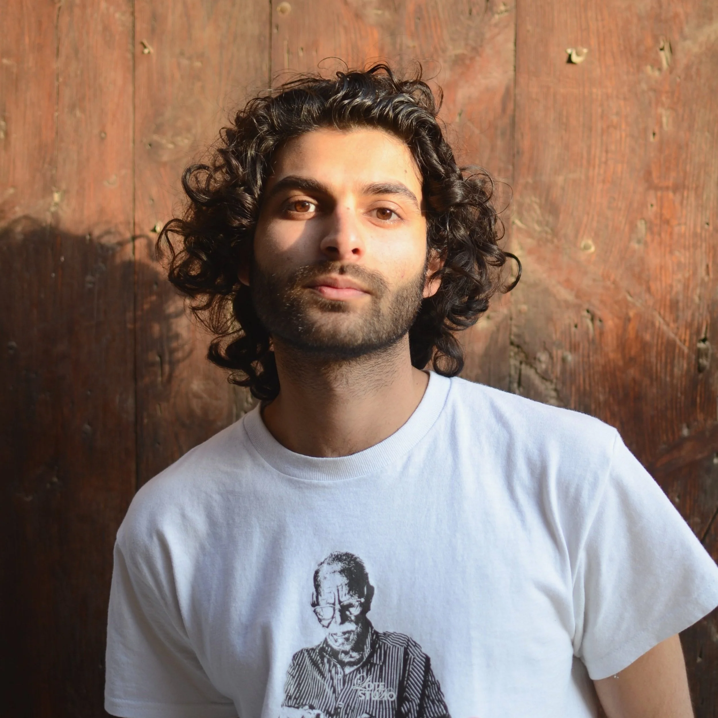 A young man with curly dark hair and a beard standing in front of a wooden wall, wearing a white T-shirt featuring a graphic of an older man with glasses.