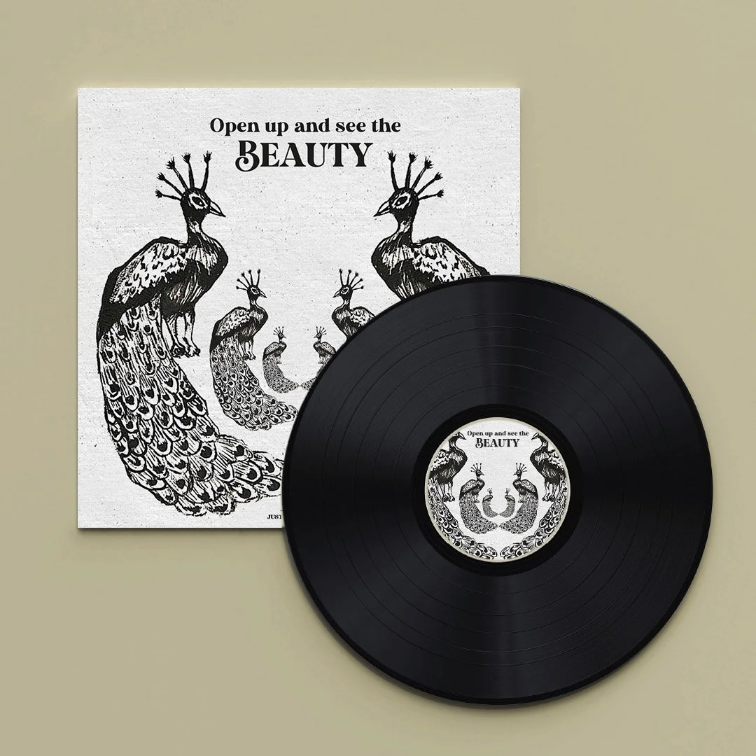 A vinyl record and cover art featuring black and white illustrations of peacocks arranged in a circular pattern with the words "Open up and see the BEAUTY" on both, placed on a beige surface.