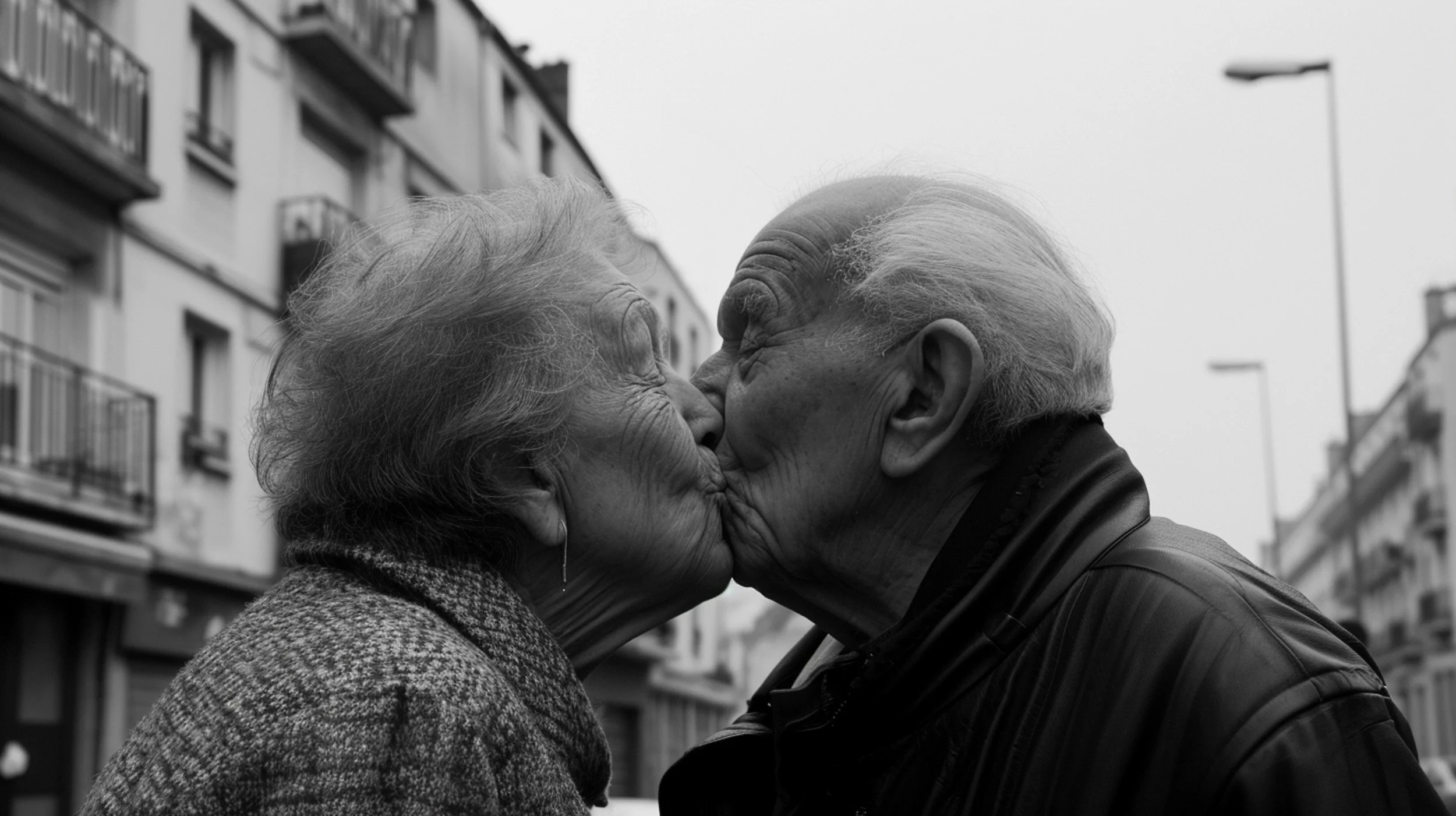 A black-and-white photo of an elderly couple kissing on the lips outdoors in an urban area with buildings and streetlights in the background.