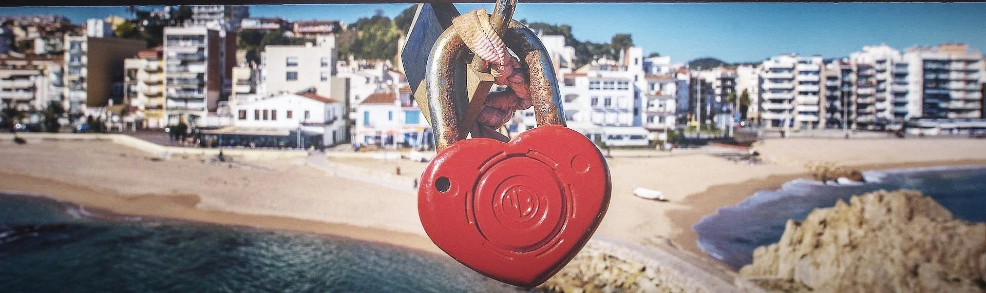 A red heart-shaped padlock attached to a metal structure overlooking a beach with buildings in the background.