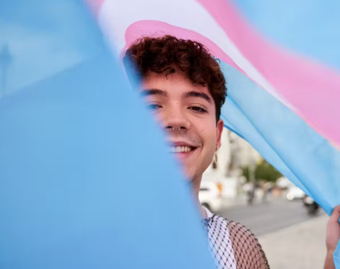 A person smiling at the camera through a transgender LGBTQ pride flag.