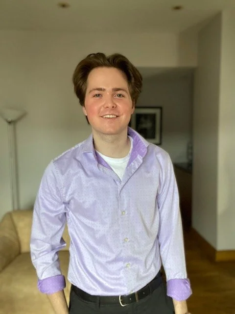 A young man with short brown hair, wearing a light purple button-up shirt with rolled-up sleeves, standing indoors in a living room.