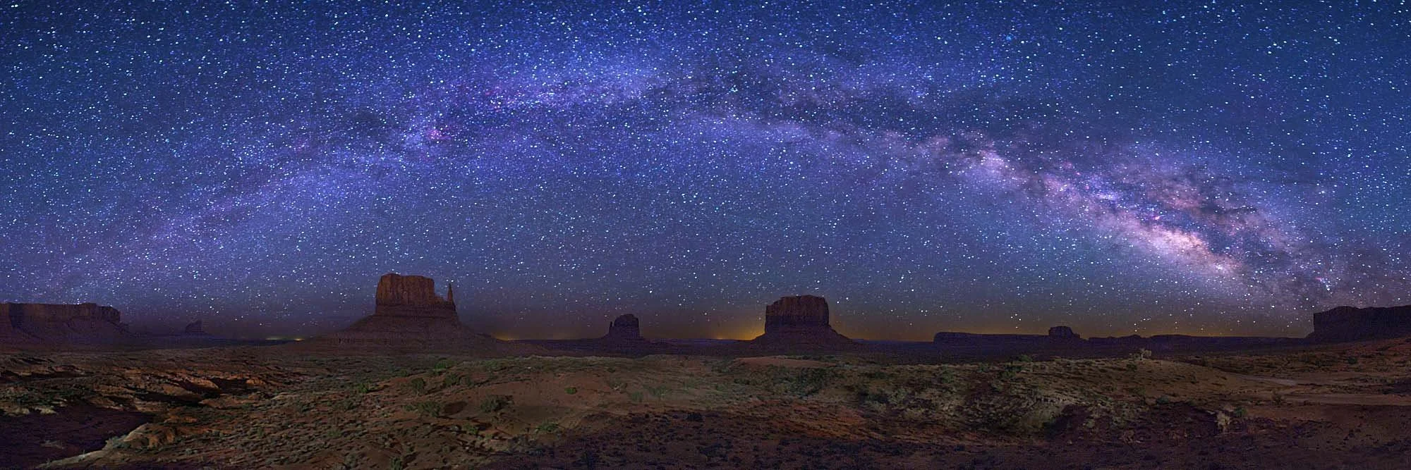 Night sky filled with stars and the Milky Way galaxy above a desert landscape with rock formations.