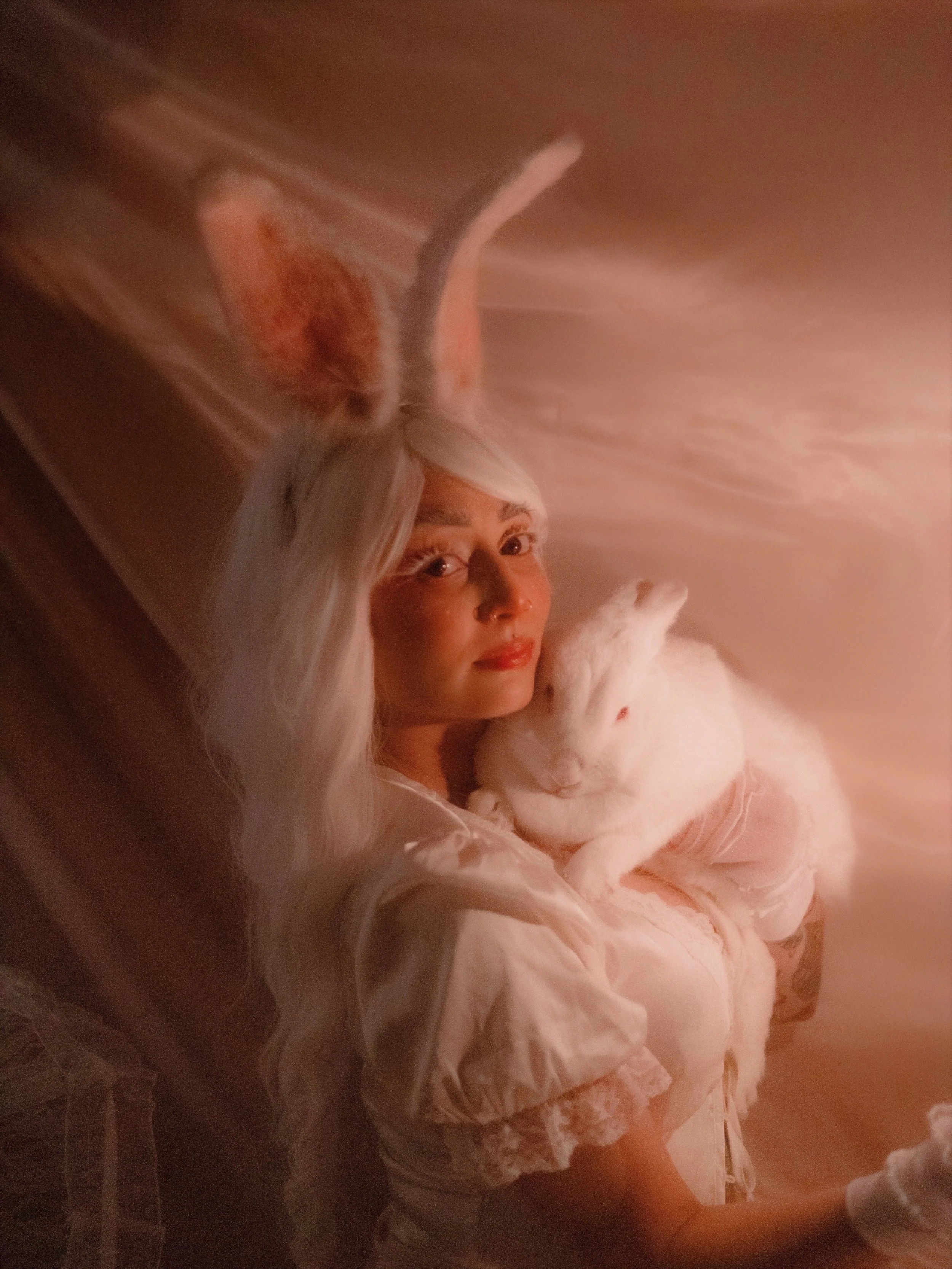 A woman with long, white hair and bunny ears holding a white rabbit in a cozy, softly lit setting.