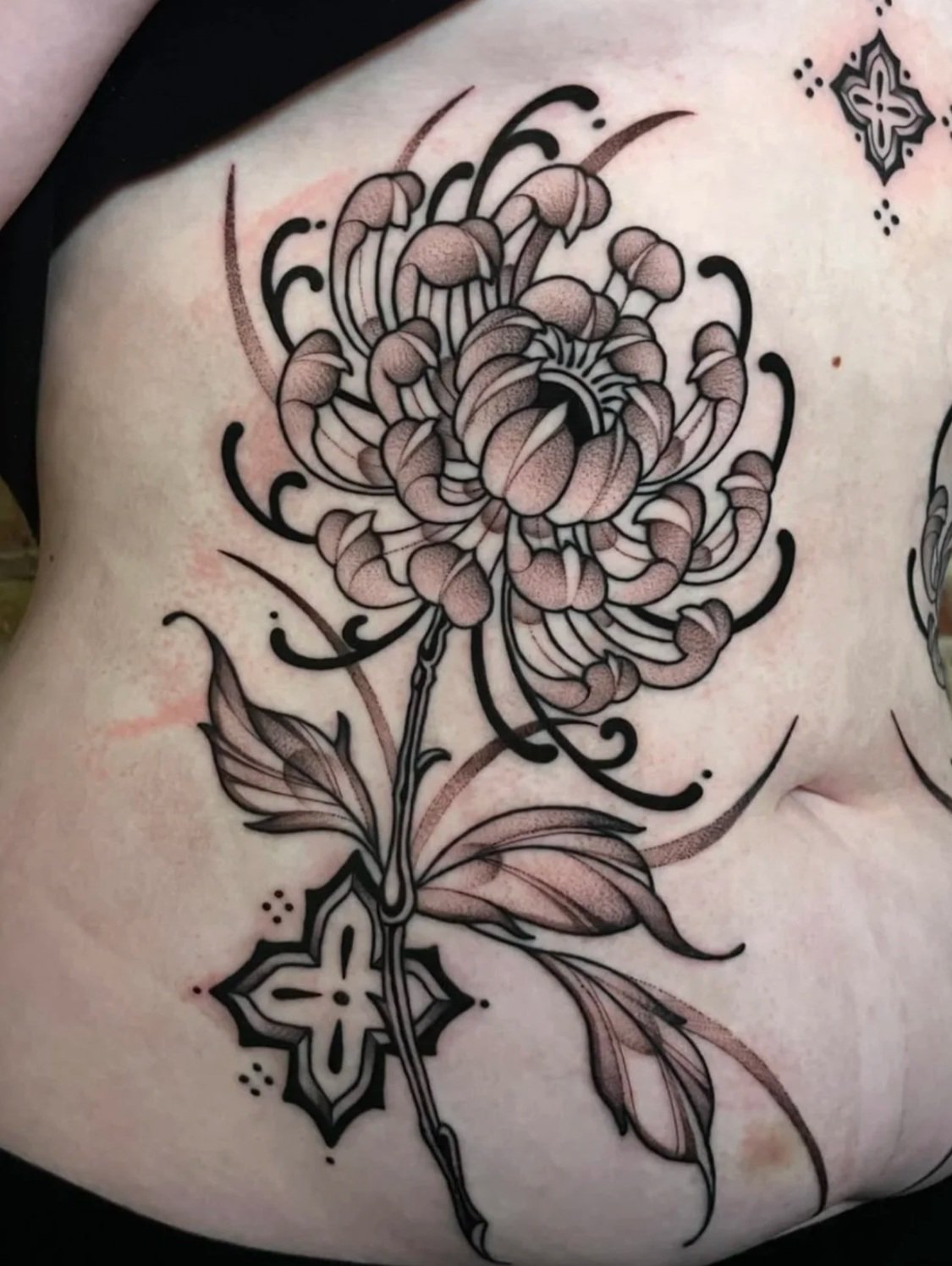 A black and gray floral tattoo featuring a chrysanthemum flower with leaves and decorative elements on a person's skin.