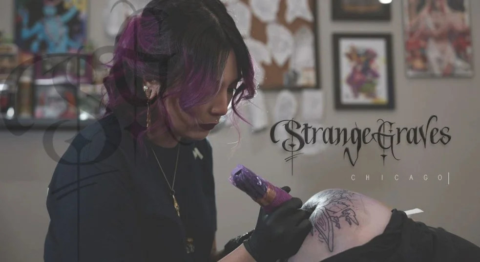 A tattoo artist with purple and black hair working on a tattoo on a client's arm in a tattoo studio. The studio wall behind them displays framed artwork and a sign that reads 'Strange Graves Chicago'.