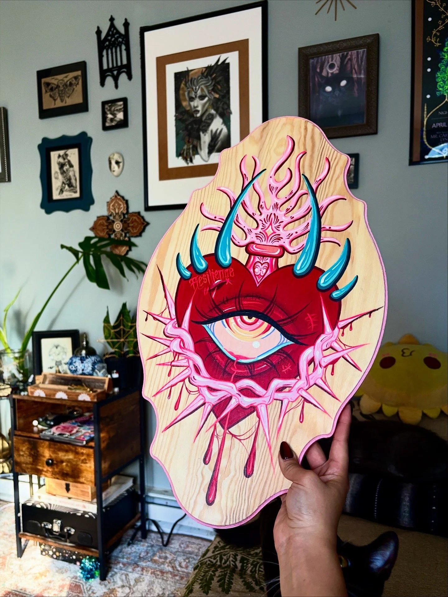 A colorful wooden artwork featuring an eye surrounded by swirling, spiked shapes and blue horns. The eye is within a red, heart-shaped design with dripping elements, set against a wood grain background.