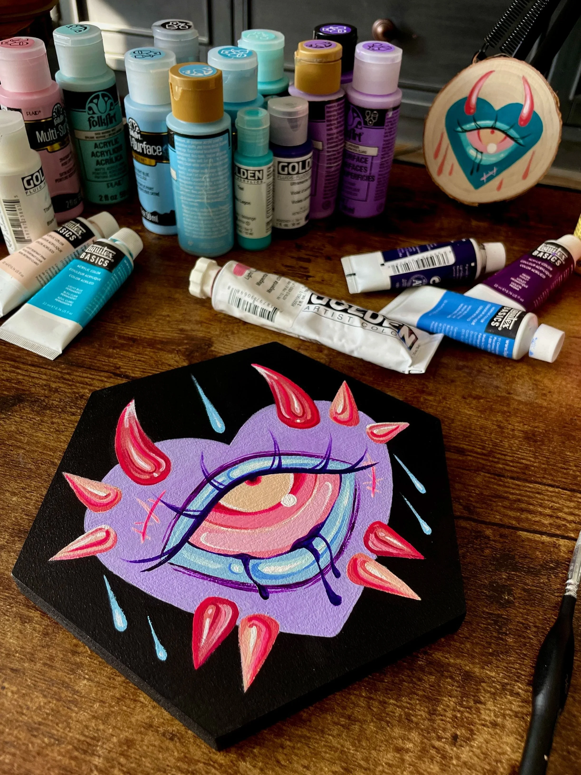 A colorful acrylic painting of a purple eye with red spikes and blue teardrops on a black hexagonal canvas, surrounded by various bottles of acrylic paint and a small circular artwork with an eye and teeth design on a wooden surface.
