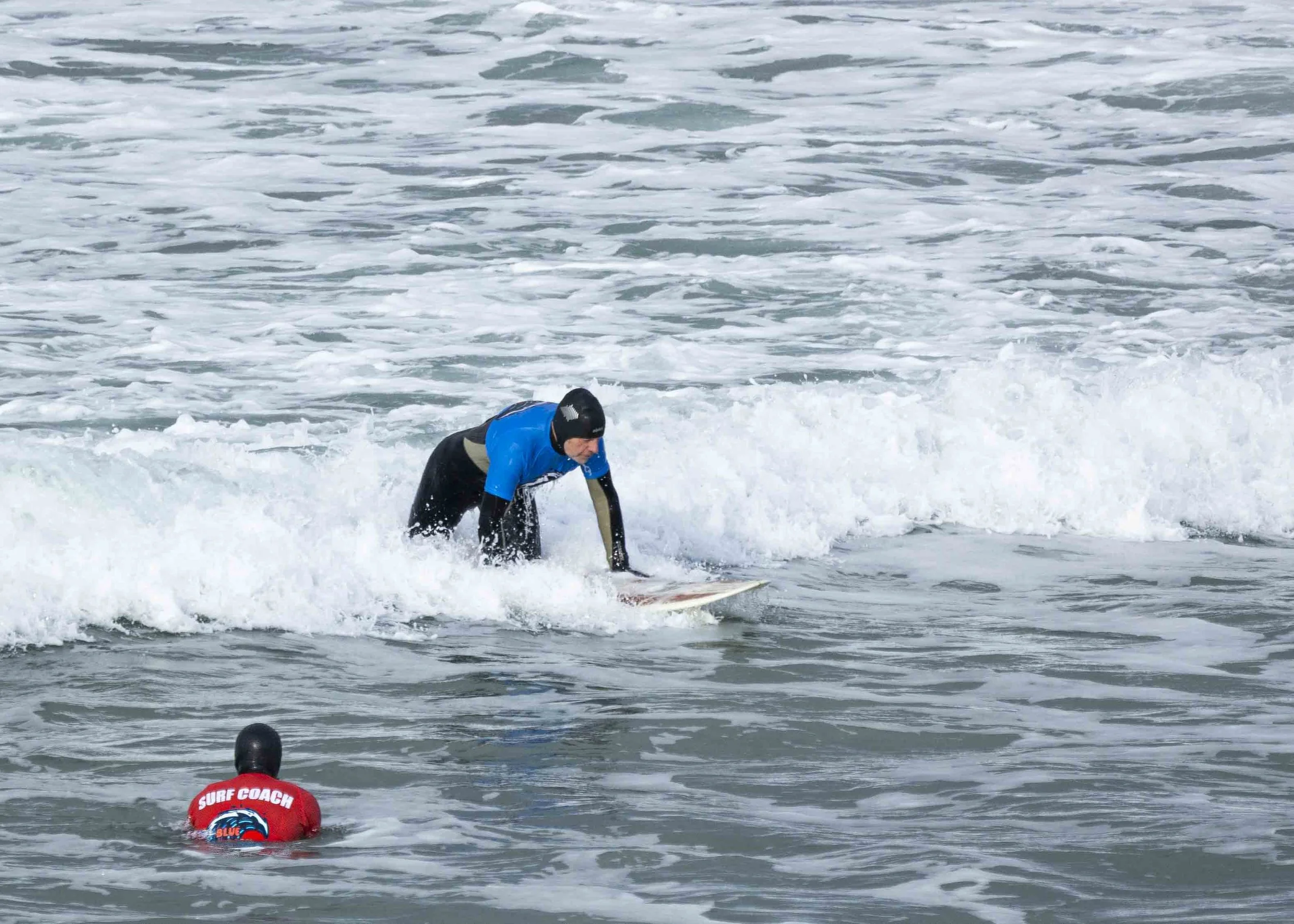 A person in a blue shirt and black wetsuit balancing on a surfboard in the ocean, with a surf coach in a red shirt and black wetsuit in the water nearby.