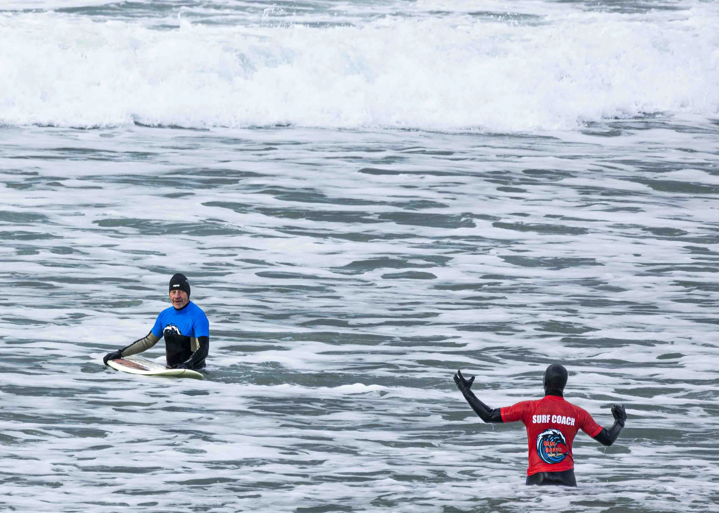 A person in a red shirt labeled 'Surf Coach' and a person in a blue shirt with a surfboard in the ocean.
