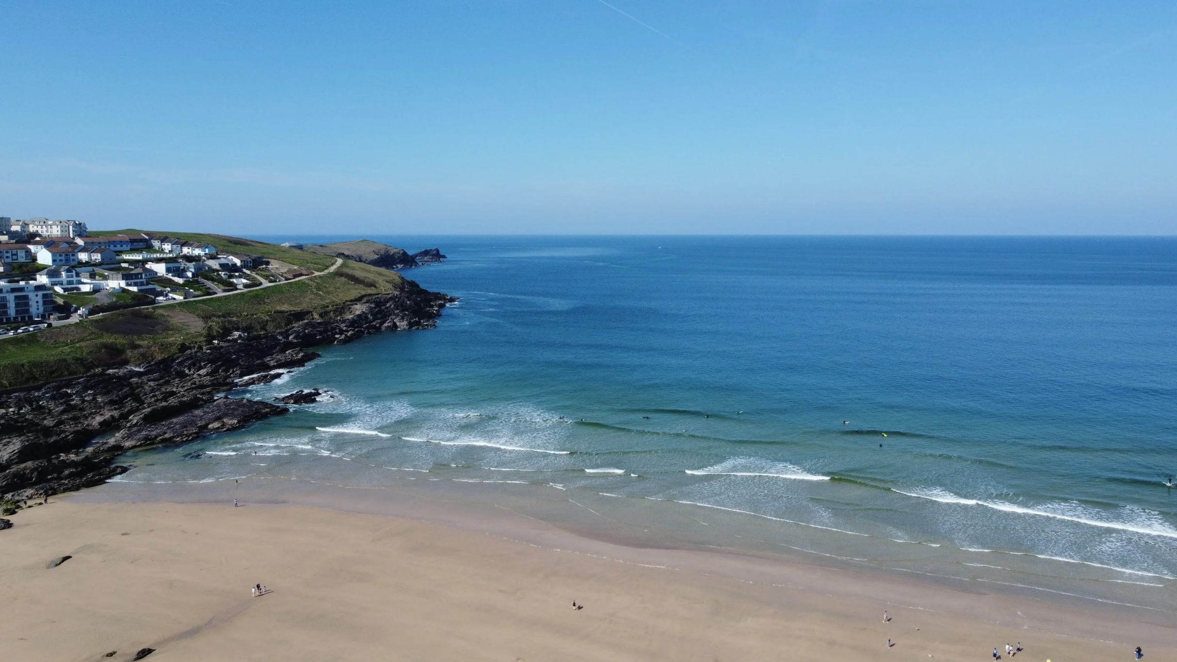 A coastal scene with sandy beach, rocky cliffs, calm blue ocean water, and residential houses on the hillside under a clear blue sky.
