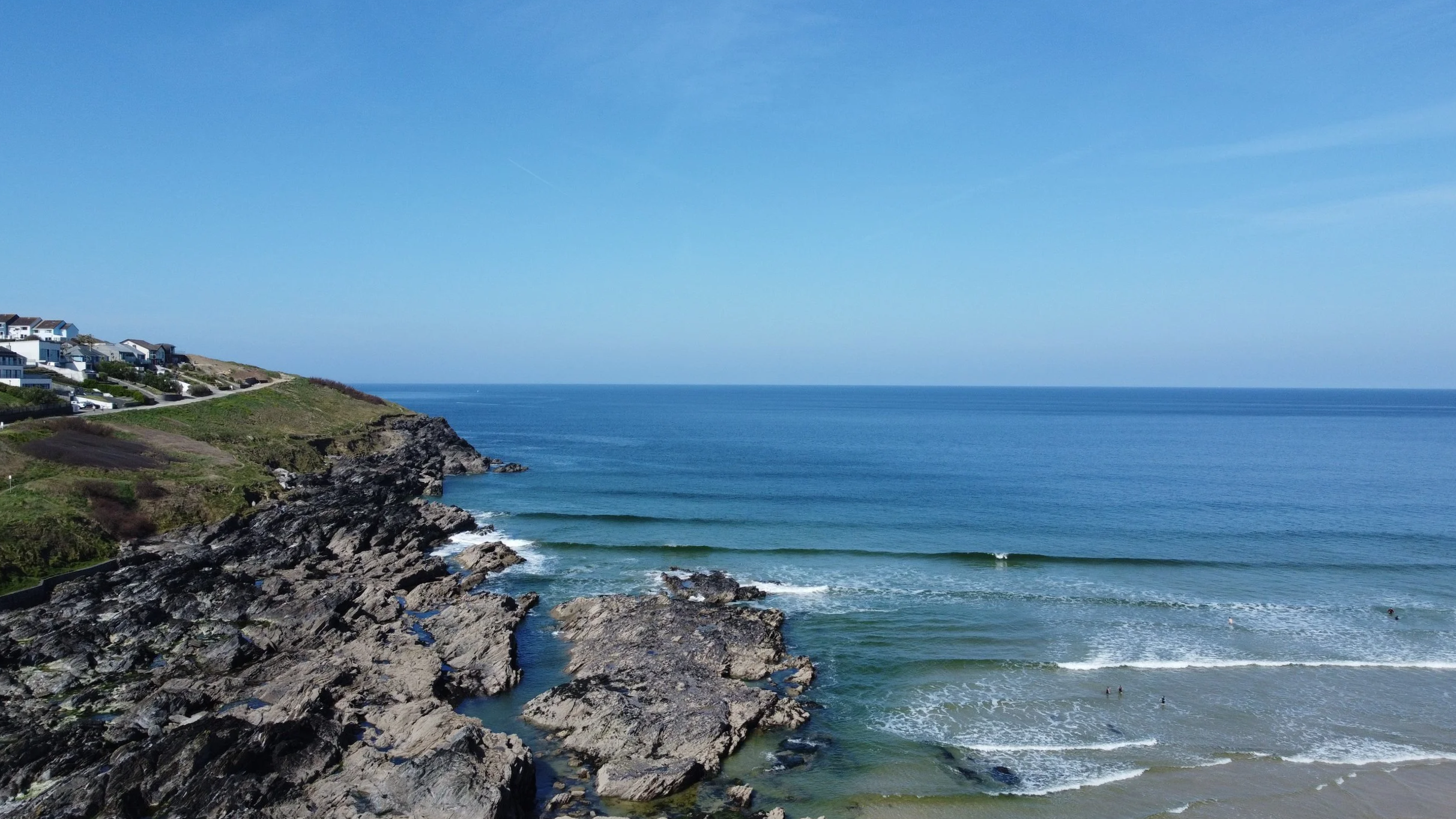 A coastal scene with rocky cliffs on the left, houses on the hill, and the ocean with small waves on the right under a clear blue sky.