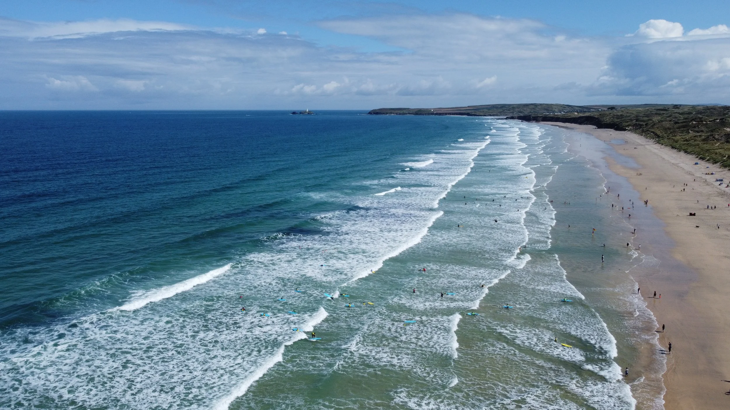 Aerial view of a sandy beach with people enjoying the shoreline, waves rolling in, and green-covered dunes in the background under a partly cloudy sky.