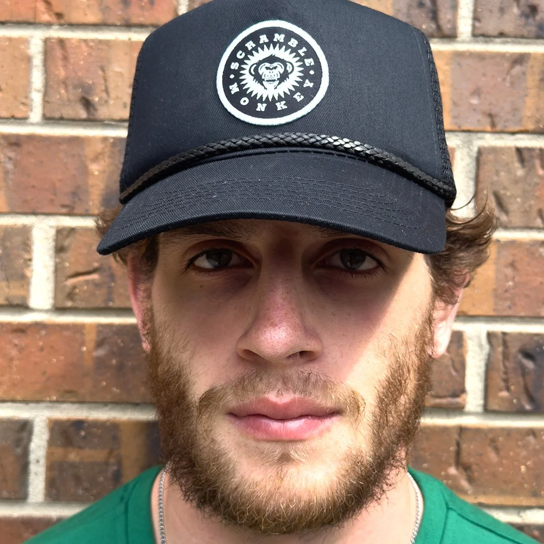 Man with beard wearing a black cap with "Scramble Monke" logo, standing in front of a brick wall.