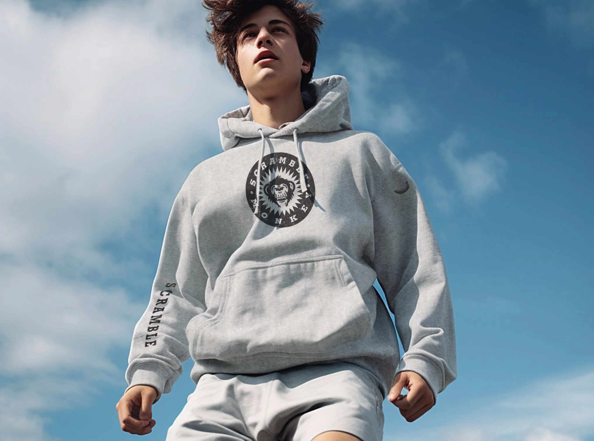 Young man wearing a gray hoodie with "Scramble" text and monkey logo on the front, blue sky background.