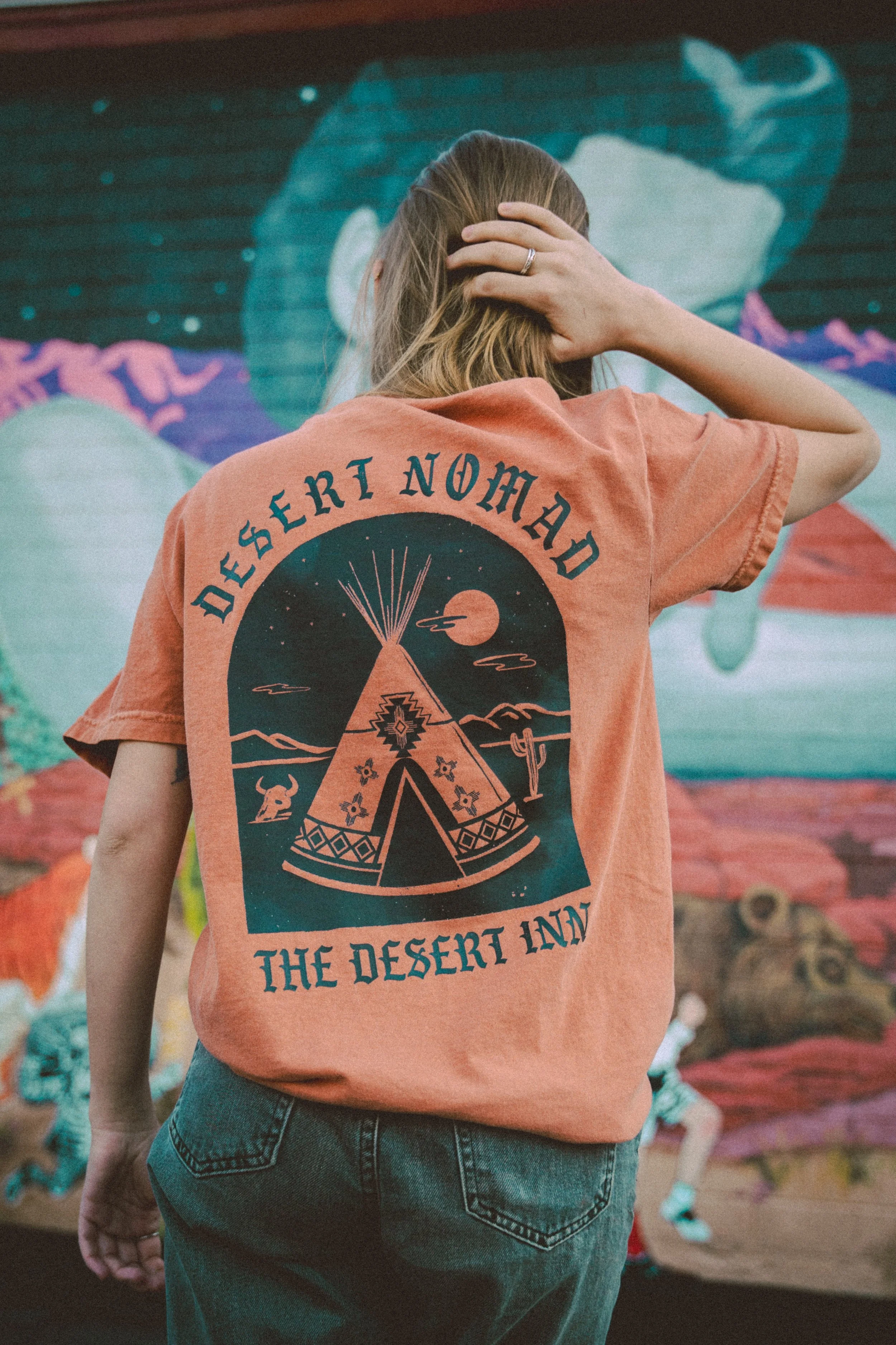 Person wearing an orange t-shirt with "Desert Nomad, The Desert Inn" text and a graphic of a teepee and desert scene, standing in front of a colorful mural.