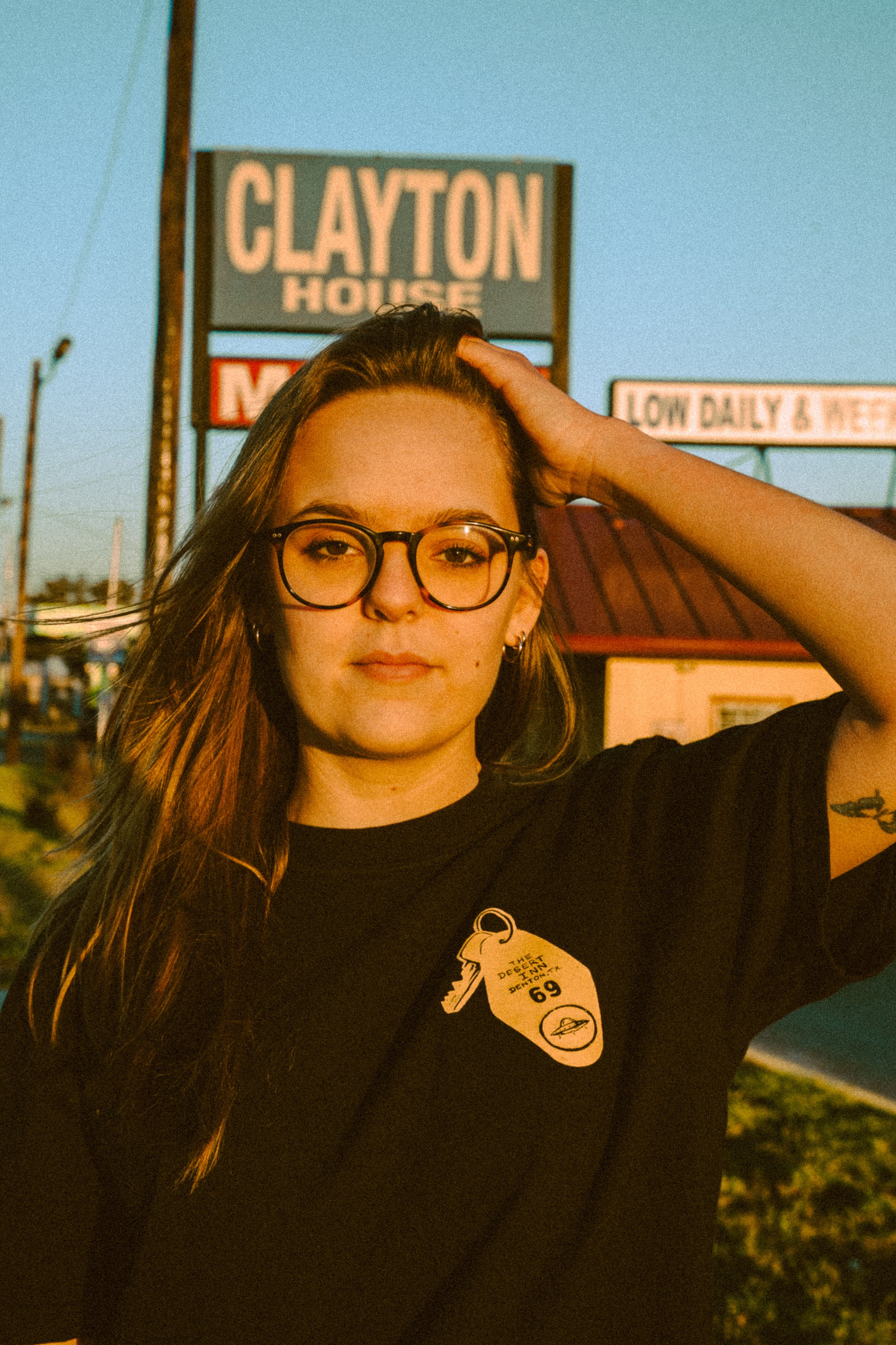 Person with glasses standing outdoors, wearing a t-shirt, with a sign in the background reading 'CLAYTON HOUSE.'