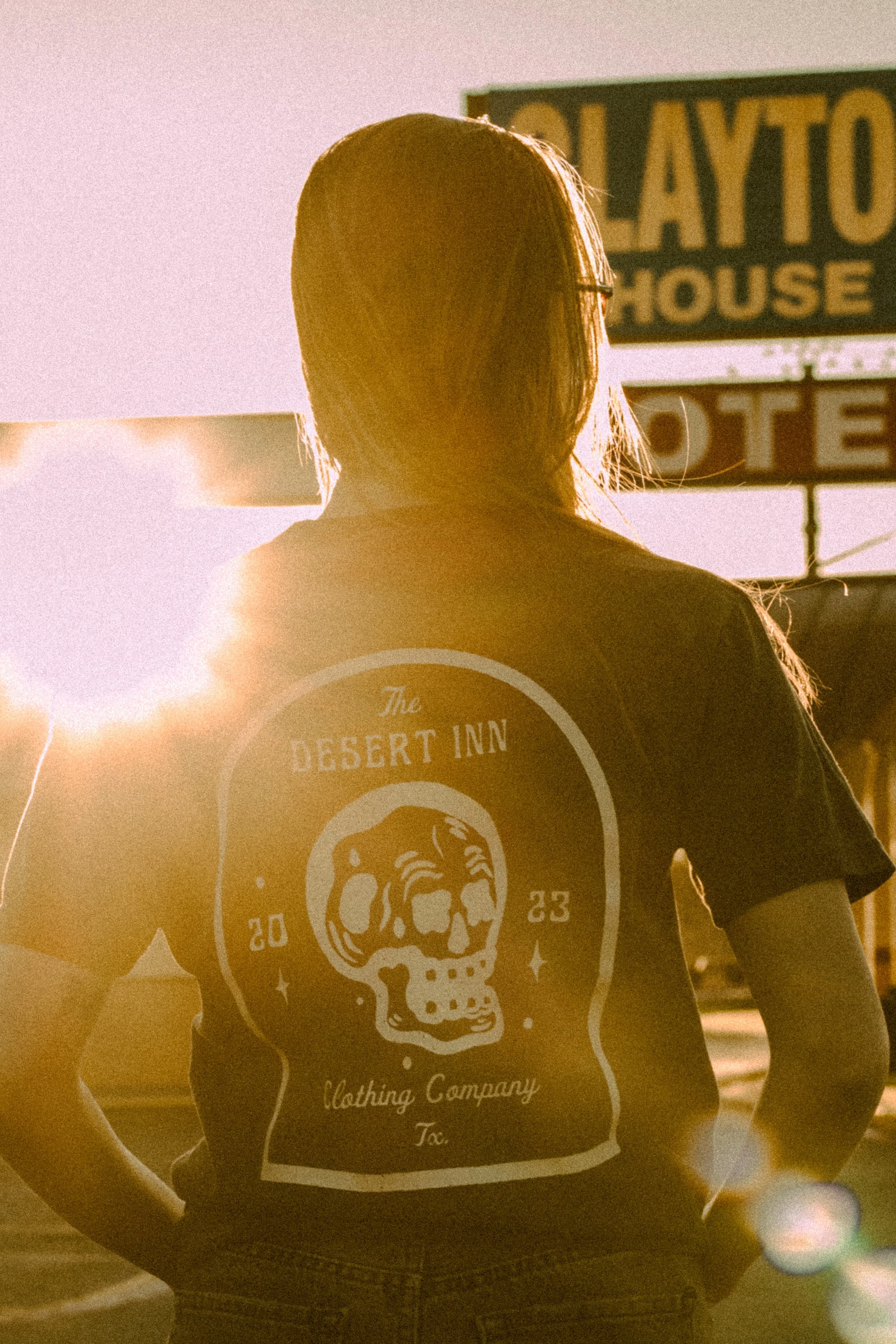 Person facing away with backlit sunlight, wearing a T-shirt featuring a skull design and text "The Desert Inn Clothing Company, TX."