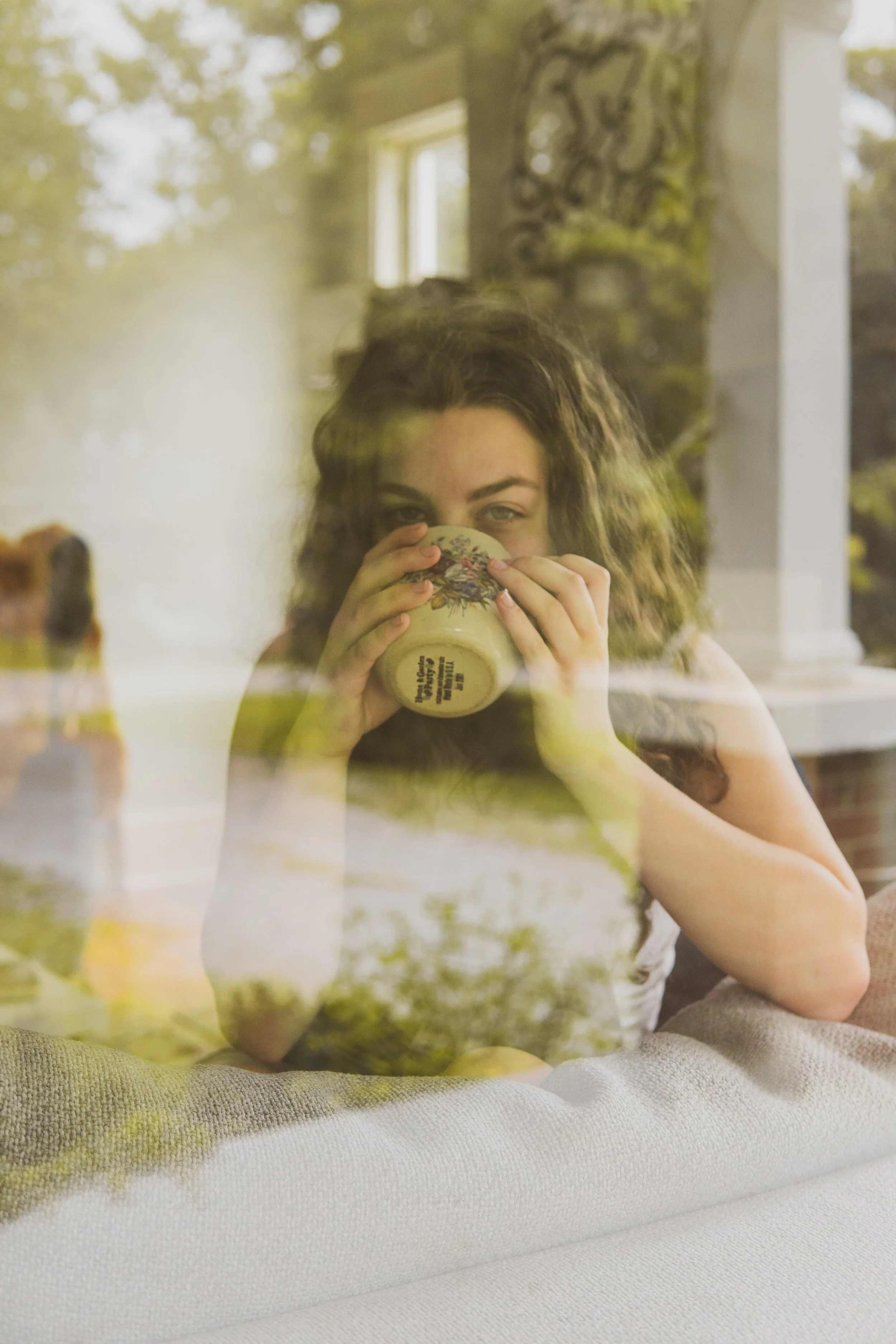 A woman sipping from a floral-patterned mug while partially obscured by reflections in a window.