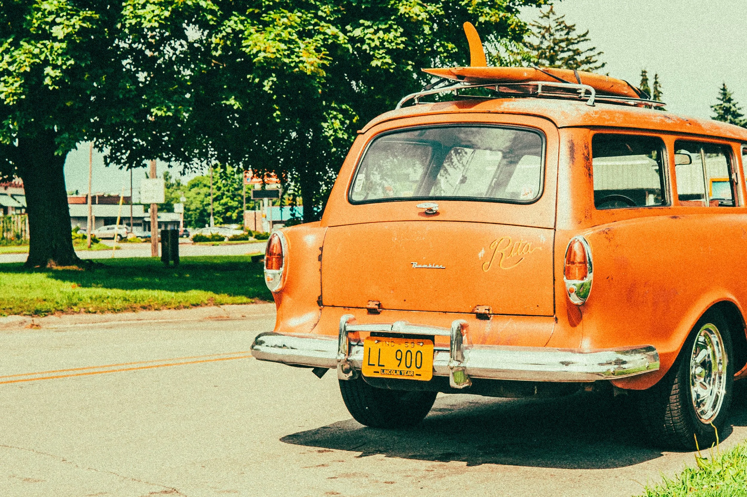 Vintage orange station wagon with a surfboard on top, parked by a tree on a sunny street.
