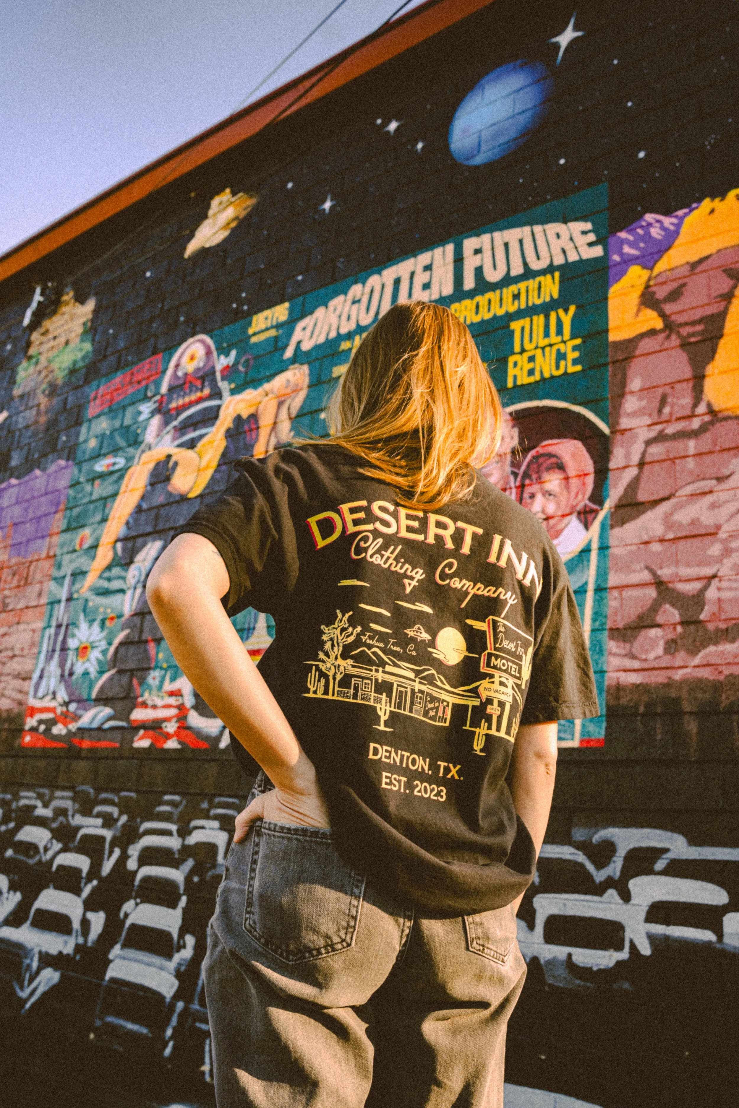 Person in a black "Desert Inn Clothing Company" T-shirt standing in front of a colorful wall mural featuring cosmic and futuristic themes.