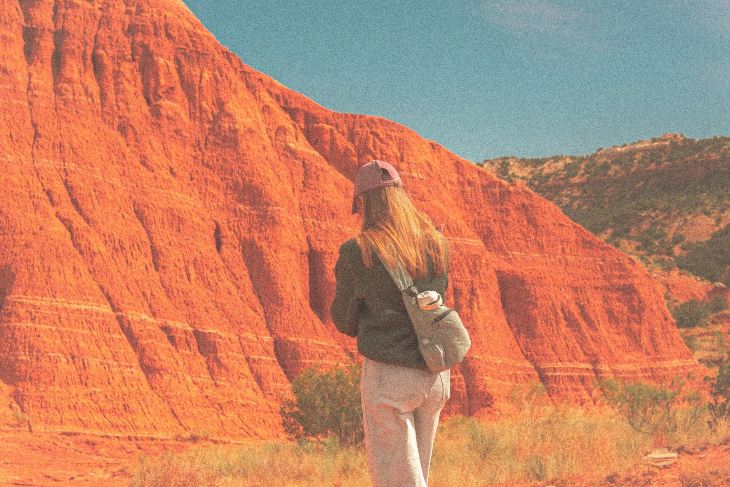 Person in cap and backpack facing red rock formations in desert landscape under blue sky.