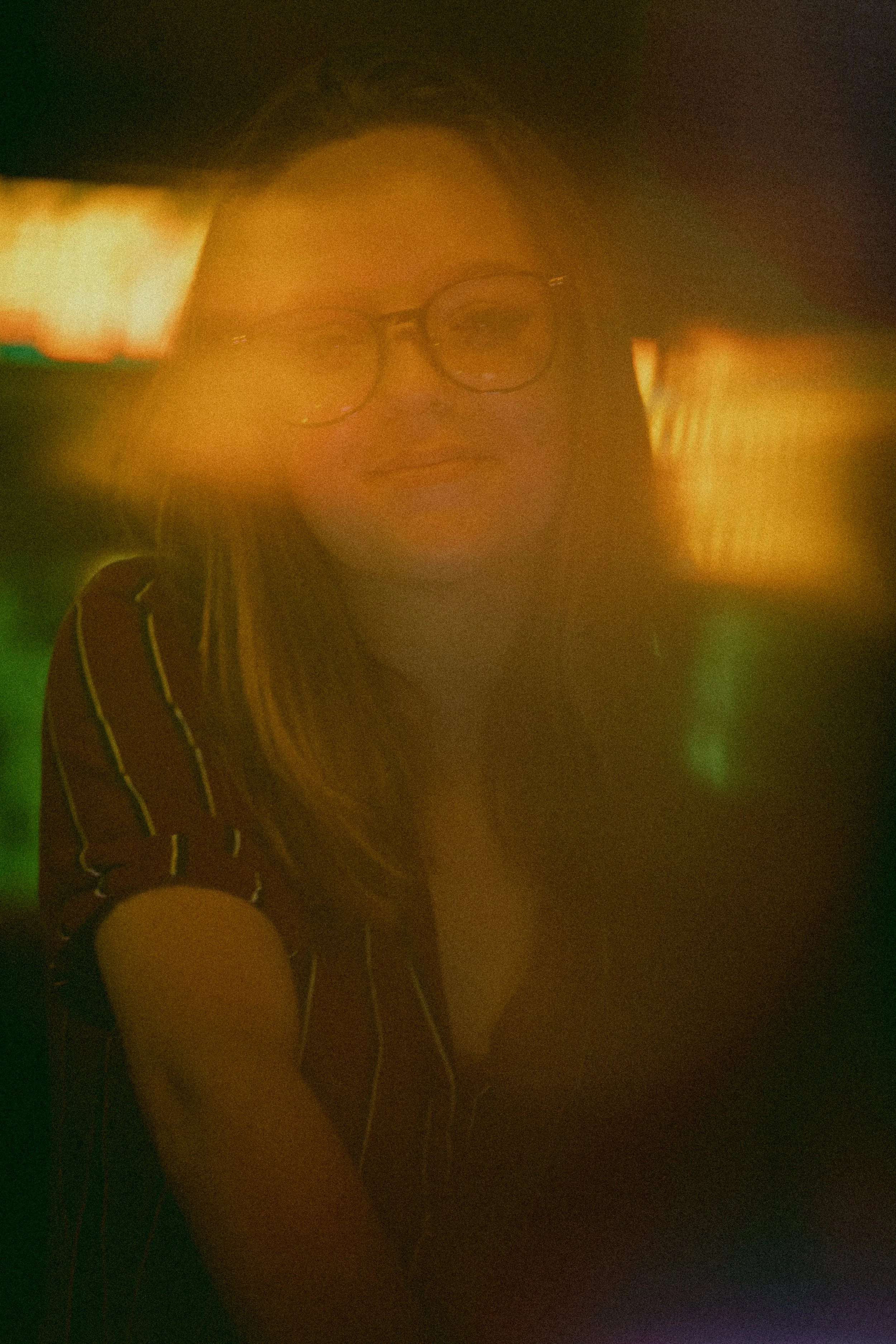 A person wearing glasses and a striped shirt standing in a dimly lit environment with a soft, warm color overlay.