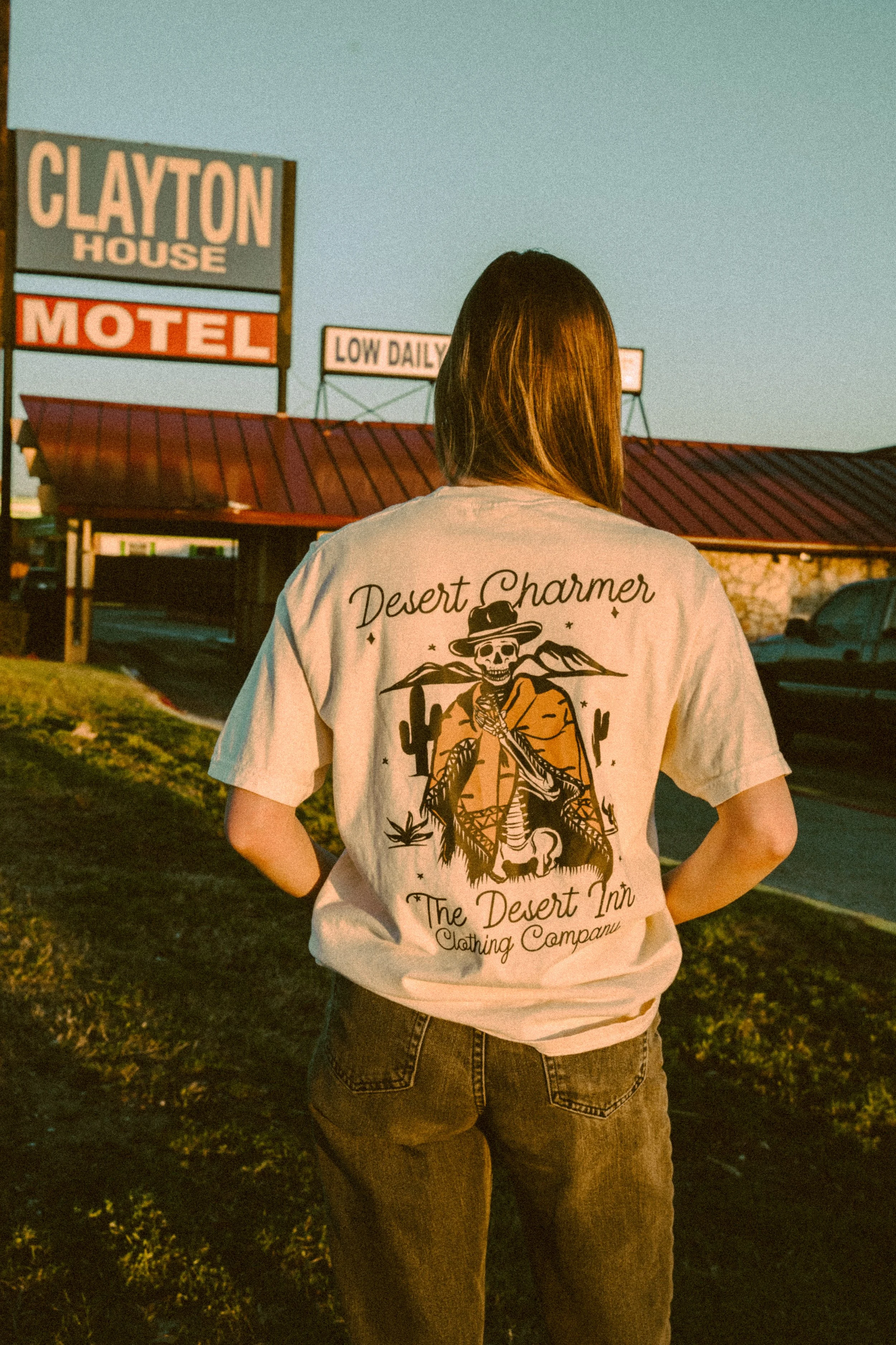 Person wearing a T-shirt with a skeleton cowboy graphic and text "Desert Charmer" and "The Desert Inn Clothing Company," standing near a motel with signage for Clayton House.